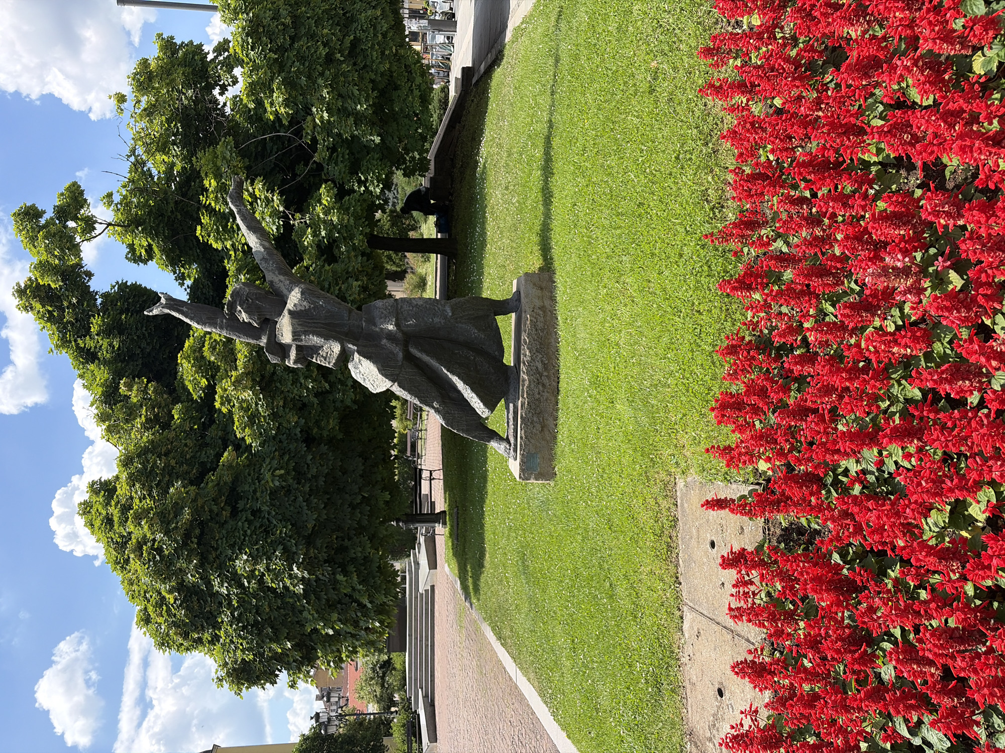 The image depicts a sunlit scene in a city park, likely in Sofia, Bulgaria, given the presence of a monument related to Bulgarian history. In the foreground, a vibrant bed of red salvia flowers lines a paved pathway. Behind the flowers, a meticulously manicured green lawn serves as a backdrop. Dominating the mid-ground is a striking bronze sculpture of a woman in dynamic motion, her arms outstretched towards the sky. The sculpture stands on a stone pedestal, itself set on the grass. Further into the background, a large, leafy tree provides ample shade, under which a lone individual is seated, possibly resting or observing the surroundings. The park features a paved walkway with benches and a tiered seating area, suggesting it is a public space designed for recreation and contemplation. The sky above is a clear blue, dotted with fluffy white clouds, indicating pleasant weather conditions. The overall atmosphere is one of peaceful urban serenity.