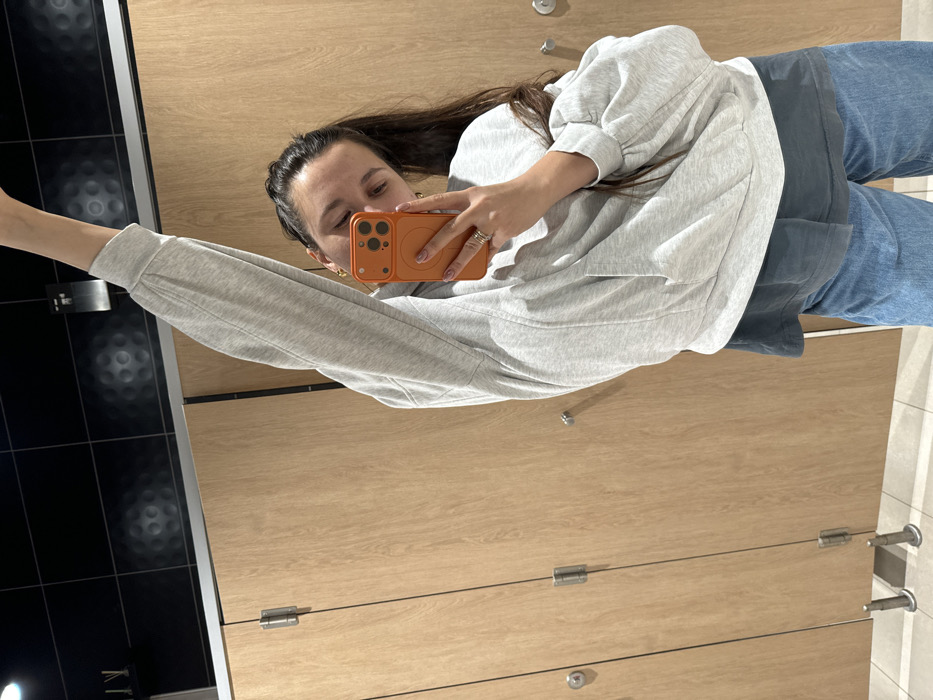 A woman takes a selfie in a public restroom. She wears a gray sweatshirt, blue pants, and a phone with an orange case. The background features wooden bathroom stalls with metal hardware and dark tiled walls. She holds the phone with her right hand, extending her left arm for the selfie. The photo is taken from an elevated angle.