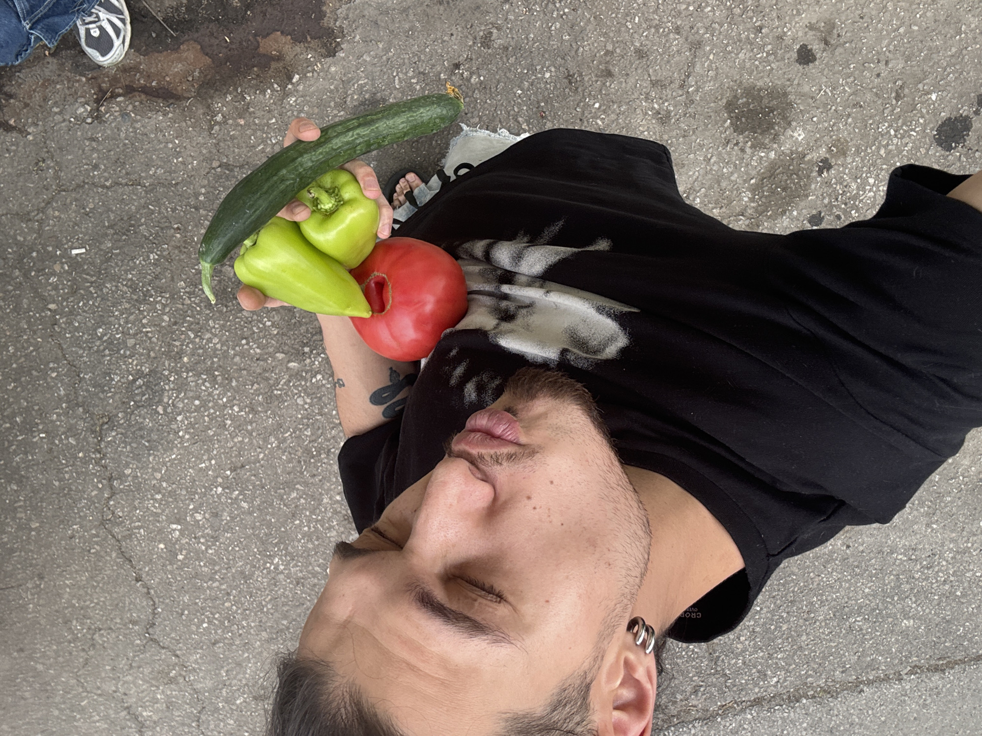 The image depicts a person lying on a paved surface, likely a sidewalk or street, in Sofia, Bulgaria. The person's head is tilted back, and their eyes are closed, with lips pursed in a kissing gesture. They are holding a collection of fresh produce: a large cucumber, two green bell peppers, and a red tomato. The person is wearing a black t-shirt with a graphic design and a silver hoop earring in their left ear. In the background, a shoe and a portion of jeans suggest the presence of another person. The ground is made of gray asphalt with visible cracks and some dark stains. The overall scene appears to be an outdoor urban environment.