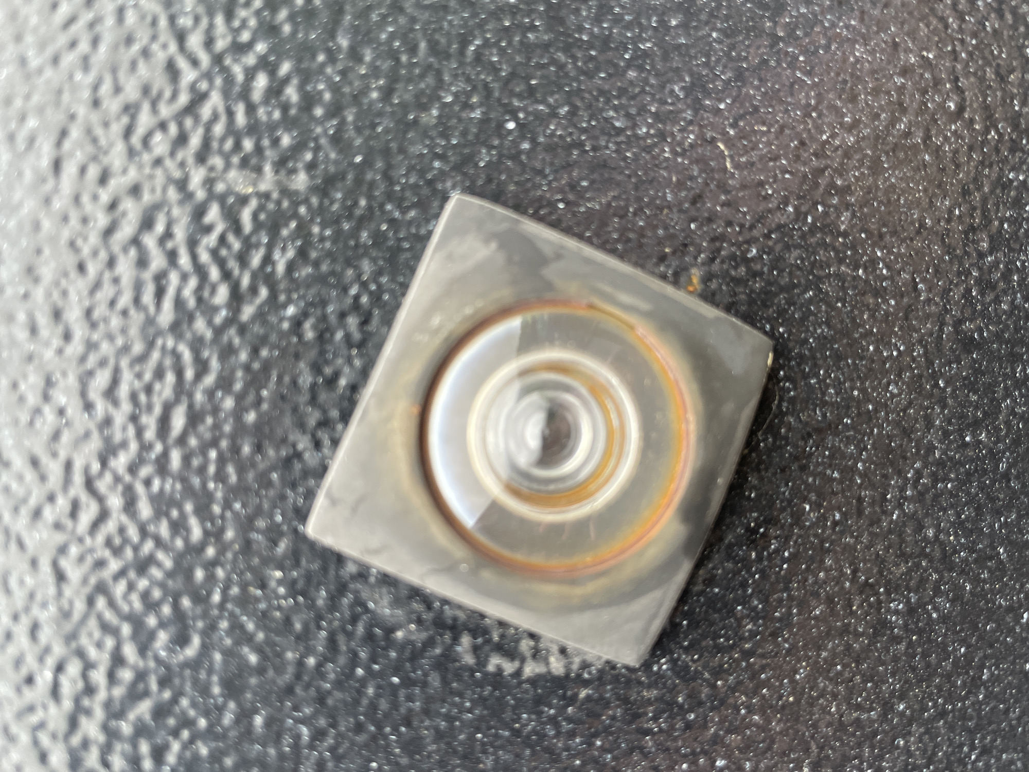 The image shows a square, metal object with a circular, lens-like structure at its center. The surrounding surface appears to be a textured, dark-colored material. There is no indication of the location being Olginka, Russia, nor any people, actions, or interactions.