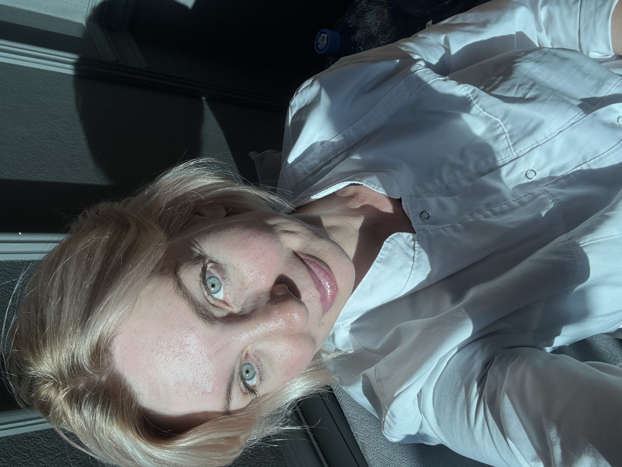 An unidentifiable woman with blonde hair and blue eyes is pictured in a close-up selfie. She is wearing a white lab coat. The lighting suggests she is near a window, with dramatic shadows cast across the scene, indicating it may be daytime. The background is dark and indistinct, with vertical lines possibly representing wall paneling or a doorframe. The overall setting is indoor, likely domestic. The location of Sochi, Russia is not evident from the image.