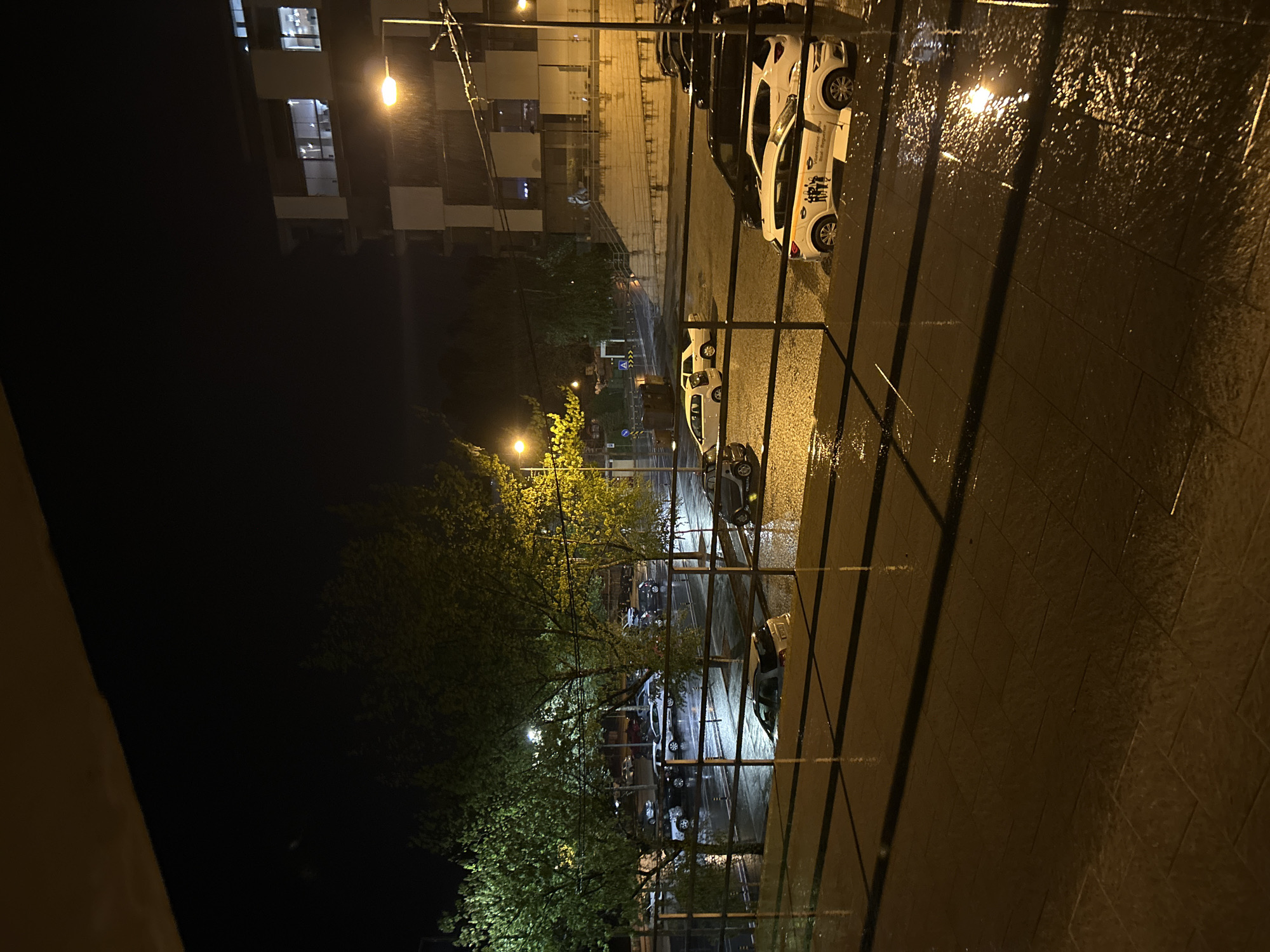 The image captures a nighttime street scene in Vila Real, Portugal, likely taken from a balcony overlooking a wet street. The foreground features a railing, with rain-slicked paving stones visible in the lower right. Across the railing, the street is also wet and reflects the light from street lamps and buildings. Several parked cars line the street, including some with distinctive white bodies and graphics, potentially indicating service vehicles. The lighting is a mix of bright, warm street lamps and the cooler light emanating from buildings in the background. The background features a building with multiple stories and lit windows, suggesting residential or commercial use. Trees with sparse leaves are scattered along the street, their branches illuminated by street lamps. The overall impression is one of a quiet, rain-soaked night in a residential or commercial area of Vila Real.