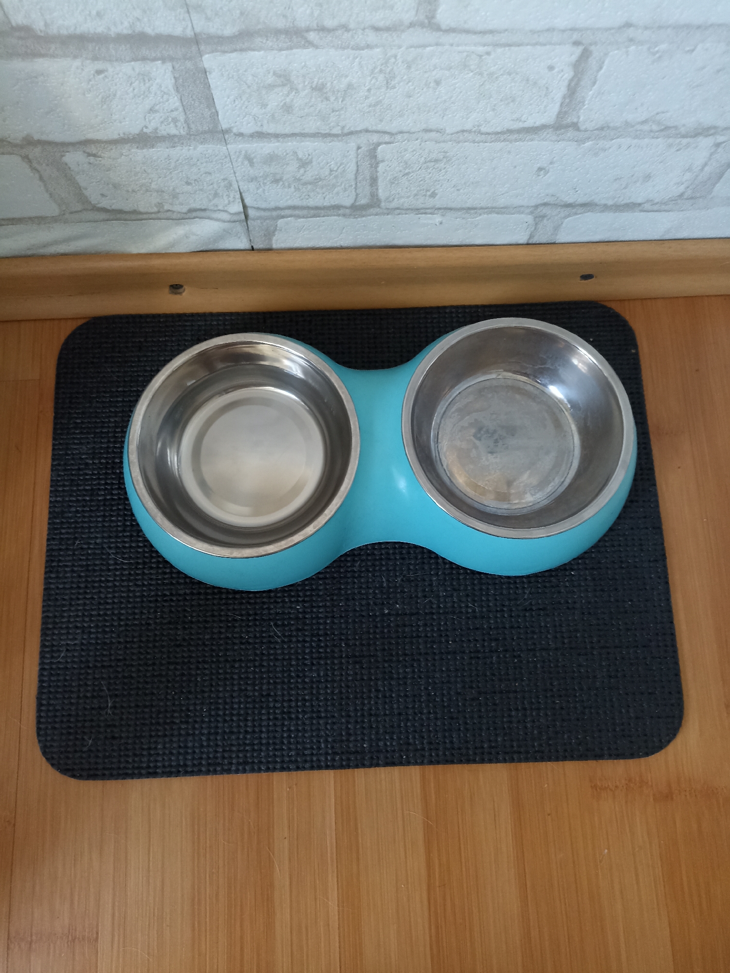 A double pet bowl set with stainless steel bowls is placed on a dark grey textured mat, which is on a light brown wooden floor. The pet bowl set is turquoise and shaped like a peanut. Behind the pet bowl set is a light grey brick-patterned wall. The image is taken from a high angle looking down at the pet bowl set.