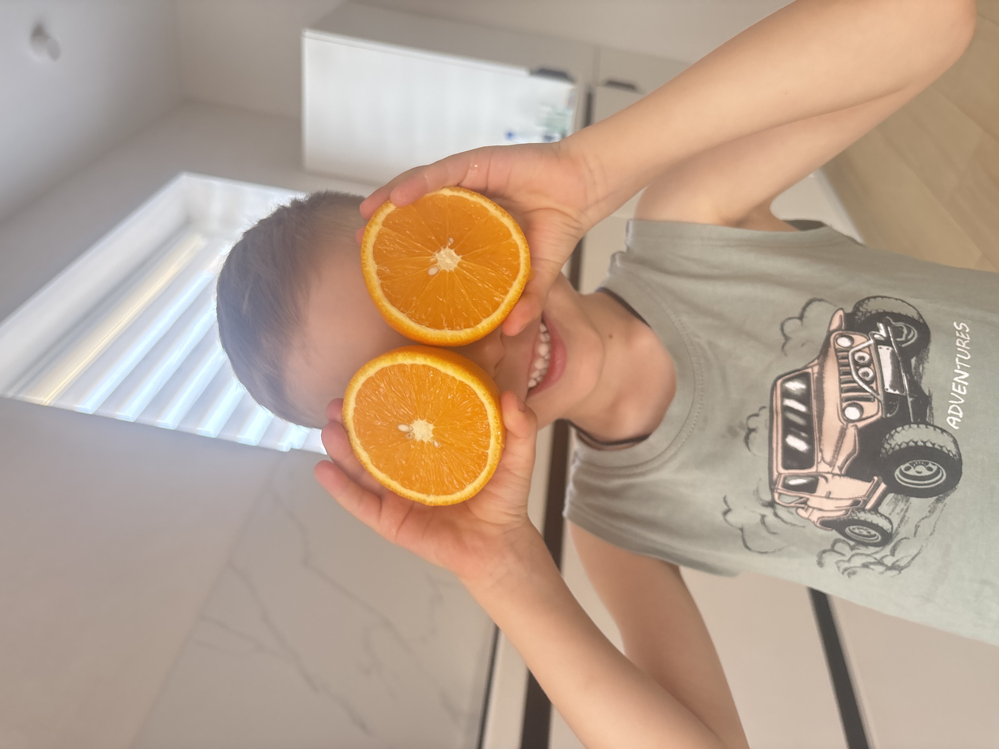 A child is holding two orange halves in front of their eyes. The child is wearing a t-shirt with a car print and smiling. The background shows a white wall and a window. The image was taken indoors. It is not possible to determine the location of Chaikovsky, Russia, from the information available in the image.