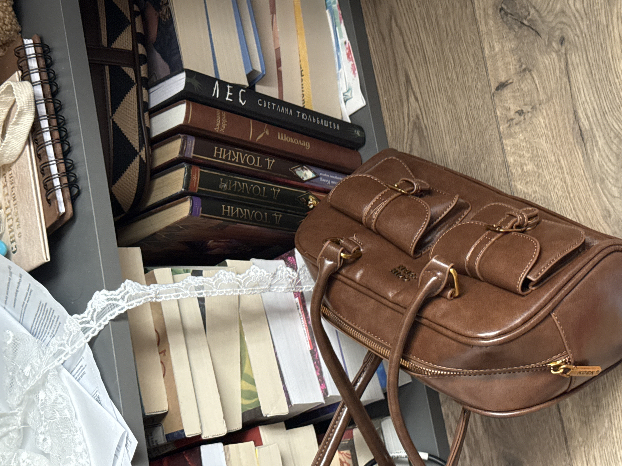 A brown purse sits on top of a bookshelf filled with books. The purse has gold hardware and a structured shape. The books on the shelf are a mix of sizes and colors, with titles in Cyrillic script. The books are stacked in an untidy manner. A white lace trim is draped over the books, and a notebook with a spiral binding is also visible. The floor is made of wood panels. Some of the books have legible titles such as "ЛЕС" (Forest), "СВЕТЛАНА ТЮЛЬБАШЕВА" (Svetlana Tyulbasheva), "Шоколад" (Chocolate), and books by "Д. ТОЛКИН" (J.R.R. Tolkien). The city of Krasnodar, Russia is not identifiable from this image.