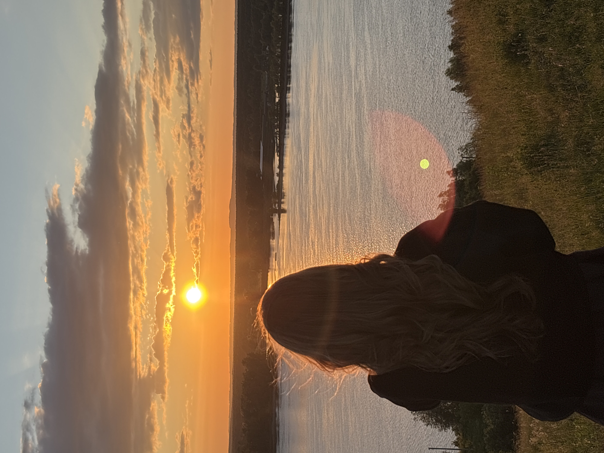 In the city of Olkhovka, Russia, a young woman with long, wavy blonde hair stands with her back to the camera, facing a vast body of water at sunset. The sun, a brilliant orange orb, is low on the horizon, casting a warm glow across the sky, which is filled with scattered clouds tinged with orange and pink hues. The water reflects the vibrant colors of the sky, creating a serene and picturesque scene. The woman is wearing a dark-colored top, and her hair is slightly windswept. She is standing on a grassy hill overlooking the water, with trees and a distant landscape visible on the opposite bank. The overall impression is one of peaceful contemplation and natural beauty.