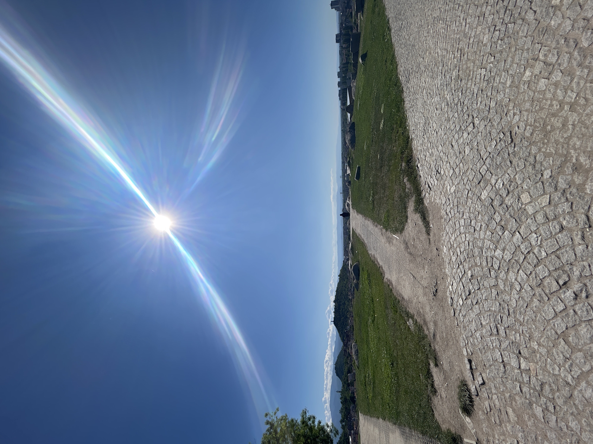 The image shows a sun-drenched outdoor scene, likely in the city of Plovdiv, Bulgaria. The sun dominates the left side of the frame, creating bright rays in the clear blue sky. The right side of the image depicts a landscape, tilted to the left. A cobblestone area is on the right, transitioning to a grassy area and a dirt path in the middle. In the distance, a city skyline is visible along the horizon. There is a hint of mountains with some clouds below it. A person is visible on the dirt path. There are some buildings in the left corner near the tree.