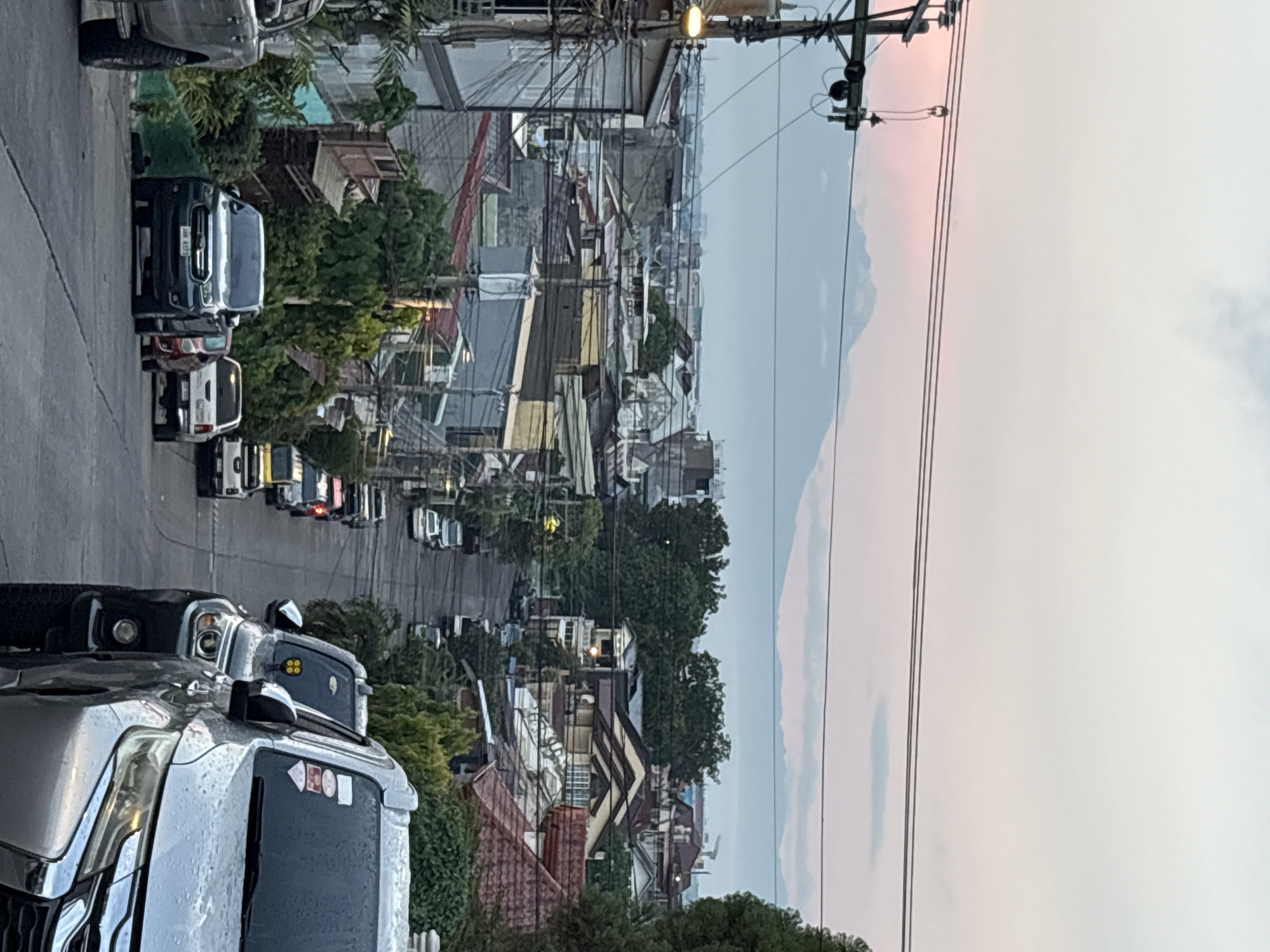 The image captures a view of a residential street in Davao City, Philippines, looking downhill towards a distant mountain range under a twilight sky. The street is lined with various houses and buildings, many with tiled roofs, indicative of a tropical climate. Numerous parked cars, mostly SUVs and pickup trucks, are visible on both sides of the asphalt road, suggesting a densely populated area. Electrical wires crisscross the frame, a common sight in Philippine urban landscapes. In the foreground, a silver SUV and a dark-colored SUV are prominently featured, both appearing wet, possibly from recent rain. The distant mountains are partially obscured by clouds and haze, with a soft pink hue in the sky suggesting the setting or rising sun. The overall scene depicts a typical urban residential environment in Davao City, characterized by its housing density, vehicle presence, and visible infrastructure, all set against a natural backdrop of a mountain.