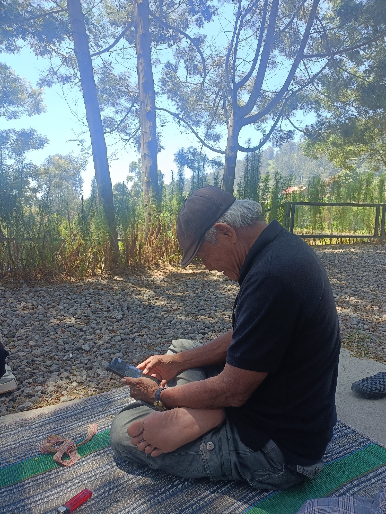 In Kopeng, Indonesia, an elderly man with graying hair, wearing a dark t-shirt and a brown cap, sits cross-legged on a woven mat outdoors. He is engrossed in looking at his smartphone, which he holds in his hands. The ground around him is covered with small gray stones, and tall trees with sparse foliage tower in the background under a clear blue sky. To the right of the man, a section of a metal fence is visible, and beyond that, rolling hills with vegetation. A red lighter and a colorful woven strap lie on the mat near the man's feet.