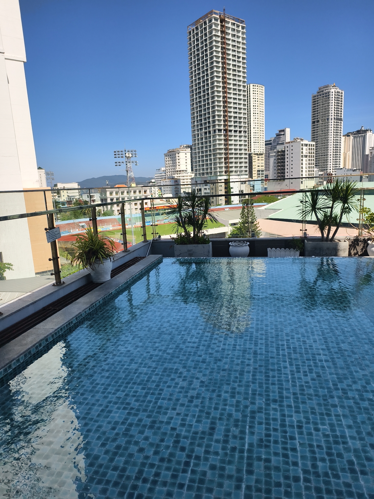 A daytime shot from a high angle shows a rooftop swimming pool in an urban area. The pool's surface is rippled, reflecting the surrounding high-rise buildings and clear blue sky. The pool is tiled with small, square blue tiles.
A glass and metal railing surrounds the pool area, behind which potted plants and greenery add a decorative touch. Beyond the railing, a cityscape is visible, with several tall buildings dominating the skyline. A sports field with stadium lighting is also visible in the middle distance. The location appears to be urban, possibly a hotel or residential building rooftop.