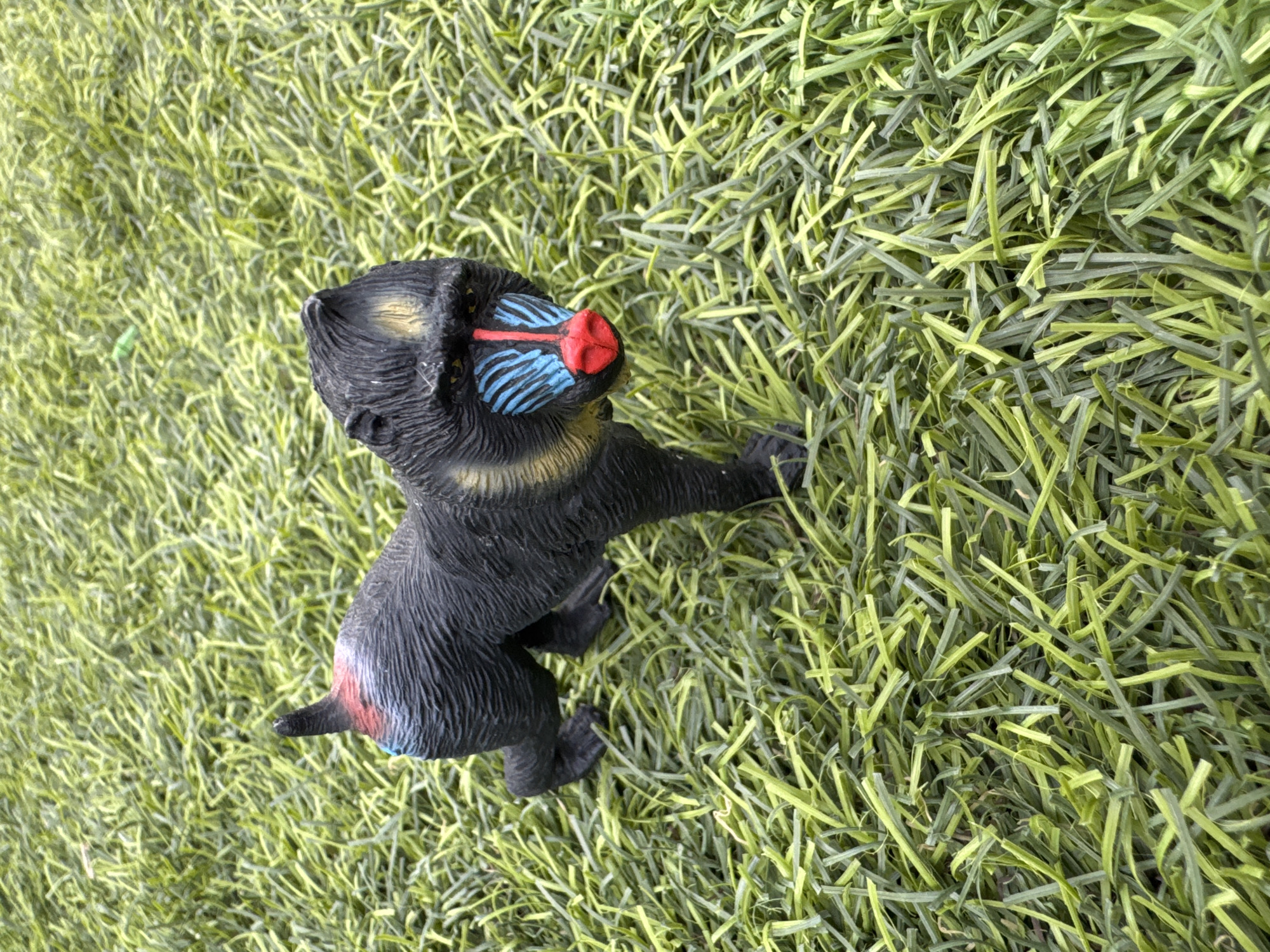 The image shows a close-up, ground-level view of a black toy baboon with a red nose and blue and yellow markings on its face, standing on artificial turf. The toy is positioned in the center of the frame, facing forward with its head turned slightly to the right. The artificial turf appears green with yellow undertones and is densely packed. The background is entirely composed of the same artificial turf, with no discernible city features or any indication of Vallada, Spain. The lighting suggests an outdoor setting, possibly during daylight hours. There are no people, actions, or interactions depicted in the scene.