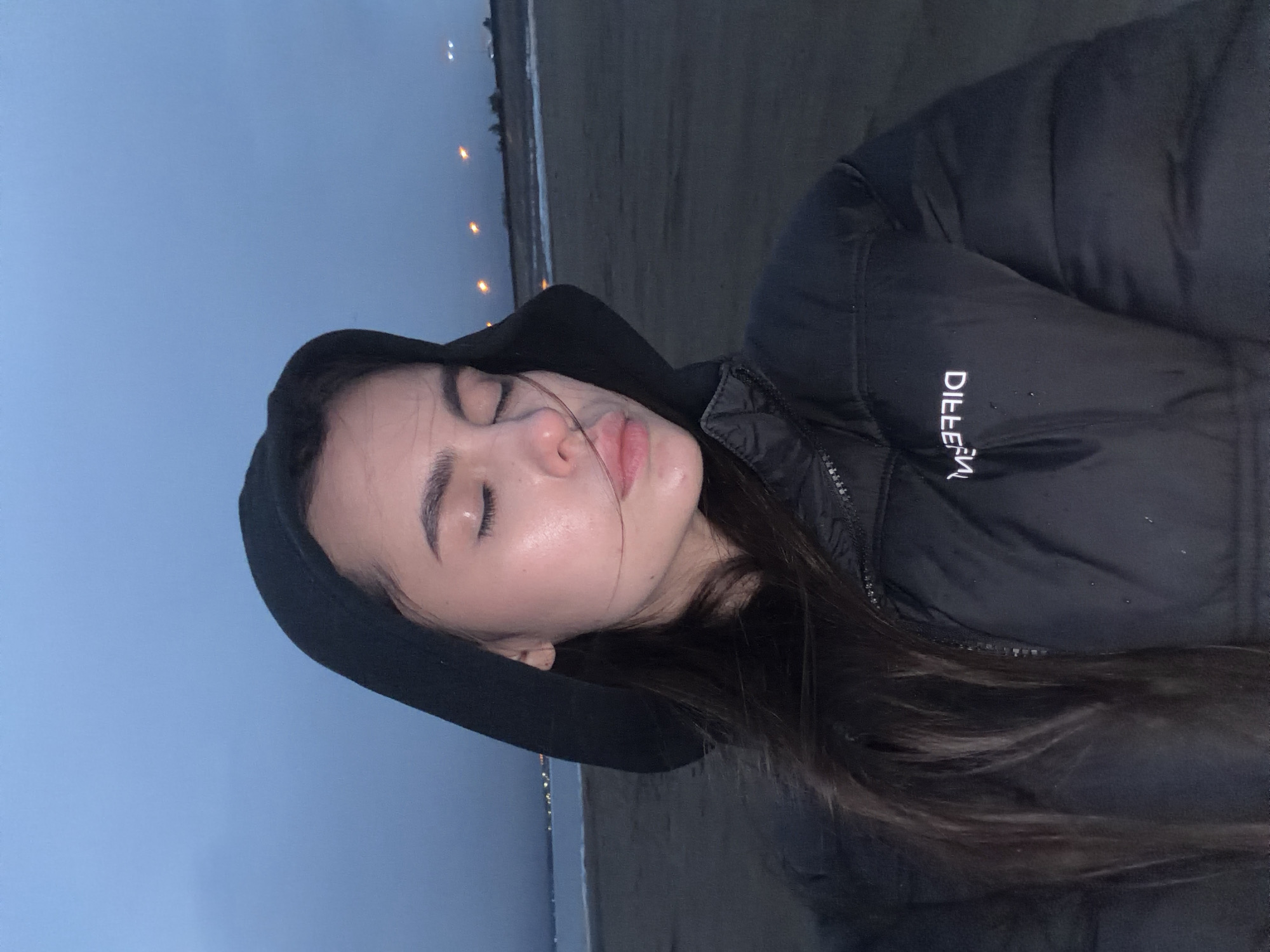 An eye-level portrait captured at dusk or twilight features a young woman with closed eyes and slightly parted lips, conveying a serene or contemplative expression. She has long, dark hair flowing over her shoulder and is wearing a black hooded puffer jacket. Her skin appears dewy or reflective.
The background shows an expansive, muted blue-grey sky above a wide, dark shoreline or beach. Across a body of water, possibly a sea or large lake, numerous small, bright lights are visible along the distant horizon, suggesting a settlement.
White, stylized text is visible on the sleeve of her black jacket. When viewed as it appears in the image, it reads "DILLEEFV", which is likely a mirrored brand name, potentially "DIFFERENT". The scene evokes a cool, calm atmosphere. The location context is Noviy, Russia.