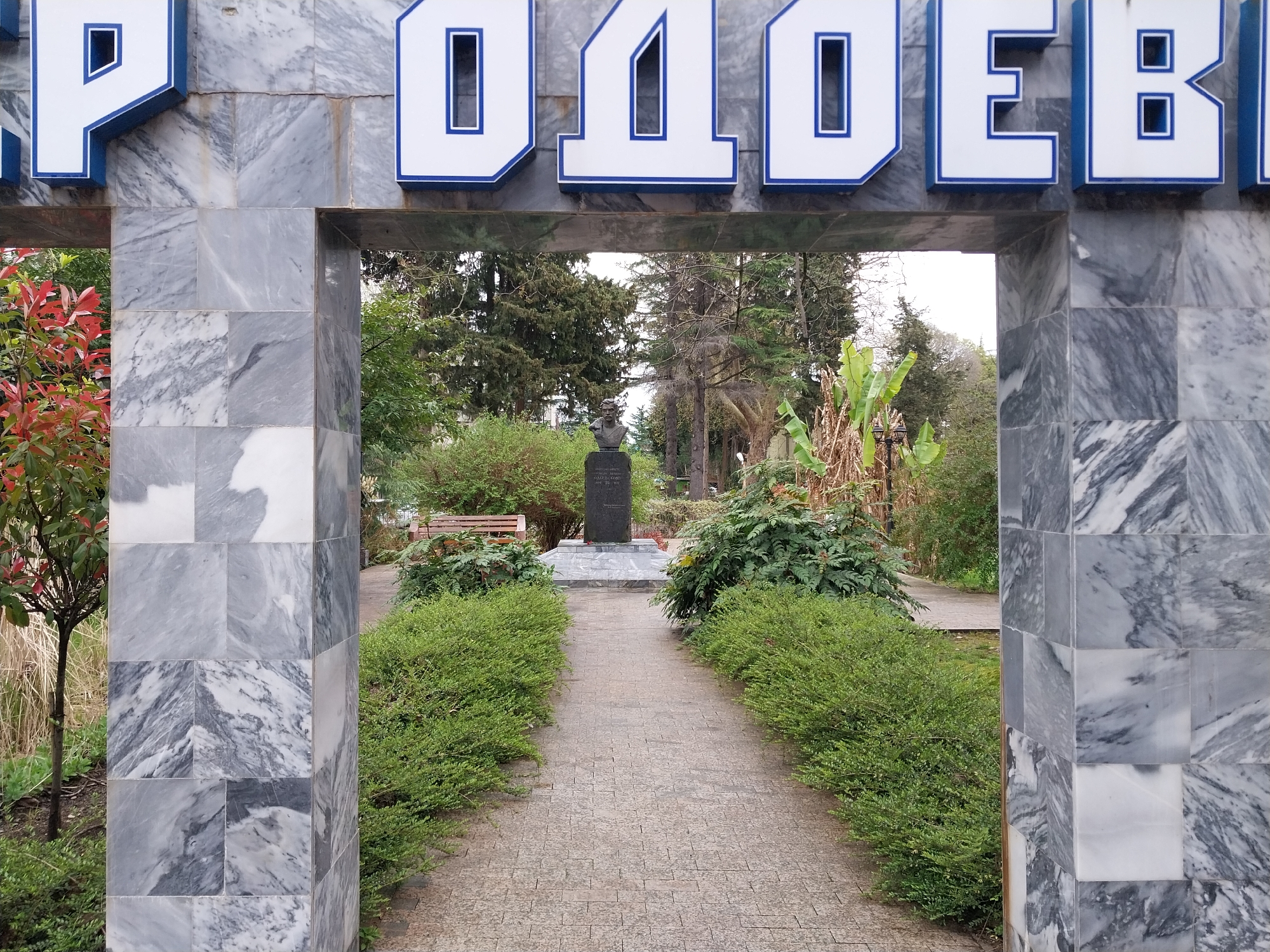 The image depicts a stone archway in a park, framing a path leading to a monument. The archway is constructed of grey and white marble, with the word "RODOEBI" in white letters with blue outlines above. The park features a paved walkway bordered by trimmed green bushes. In the distance, a dark bust stands atop a stone pedestal, surrounded by greenery and trees. A wooden park bench is visible to the left of the monument. The location is Sochi, Russia.
