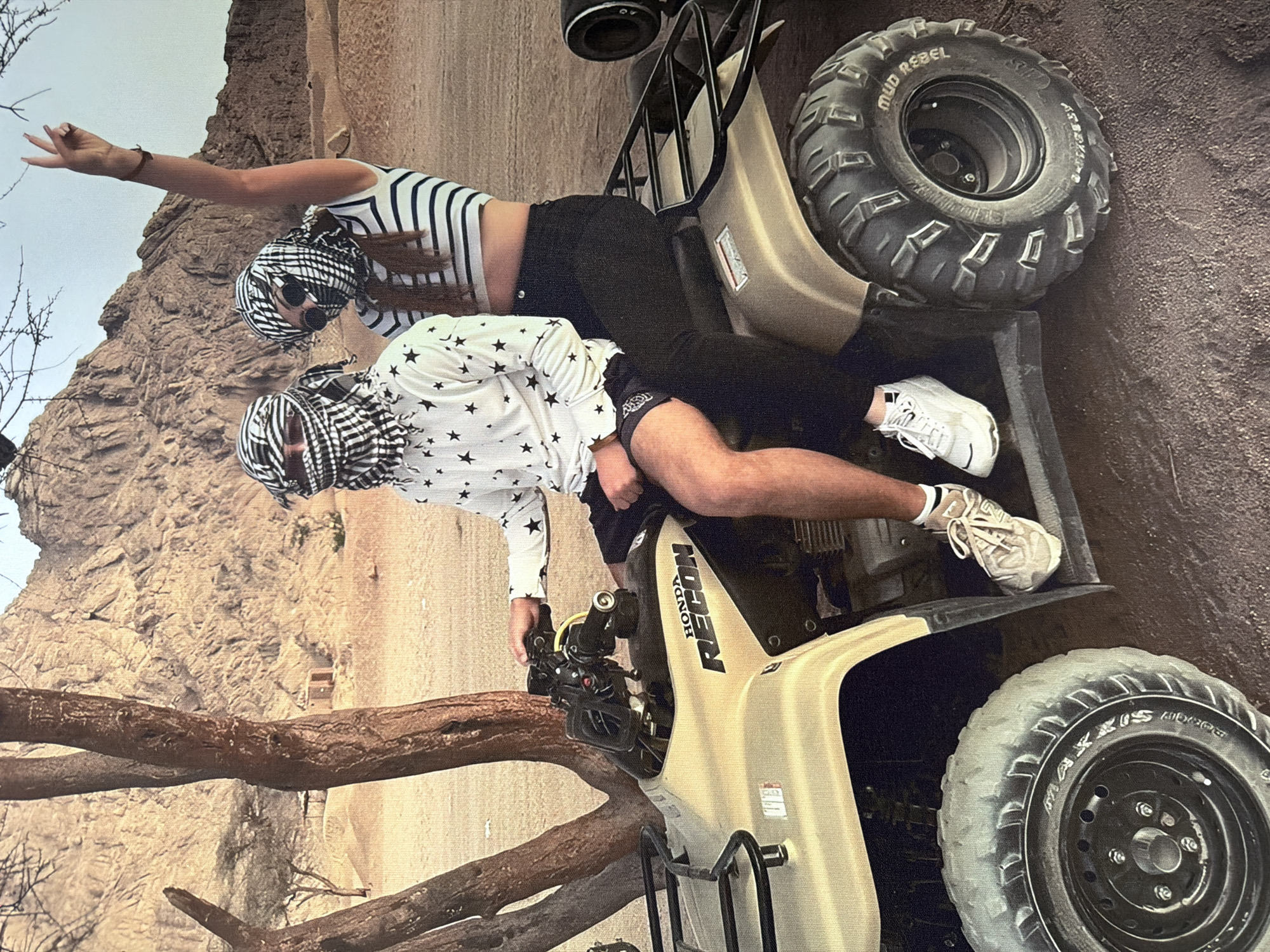Two individuals, one male and one female, are seated on a tan Honda Recon ATV in a desert-like environment with rocky mountains in the background. Both are wearing keffiyeh headscarves. The male, on the left, is wearing a white shirt with black stars and dark shorts. The female, on the right, is wearing a striped crop top and black pants, with her right arm raised in a peace sign. The ATV has black tires with "MUD REBEL" and other lettering visible on the rear tire. The ground is sandy and reddish-brown. The image does not depict the city of Strășeni, Moldova.