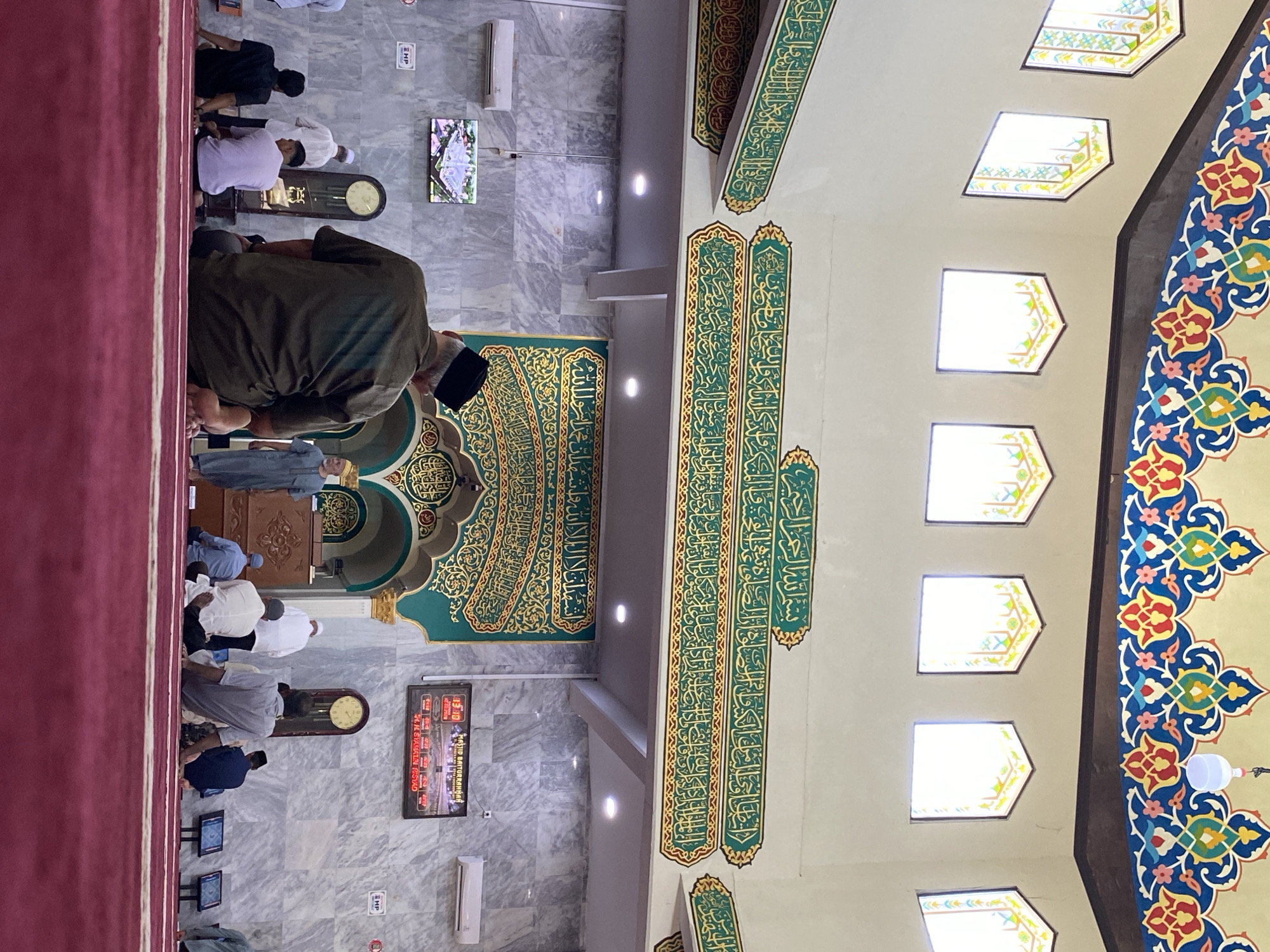 This image depicts the interior of Masjid Baiturrahman in Lhokseumawe, Indonesia. The mosque features a high ceiling adorned with colorful, ornate Arabic calligraphy and geometric patterns. Several arched windows with stained glass are visible along the upper walls, letting in natural light. The walls are clad in marble with a gray and white veined pattern.
In the foreground, several men are seated on a red carpeted floor, facing towards the front of the mosque. One man, wearing a dark green shirt and a black cap, is in the center foreground, seen from behind. Other men are dressed in various light-colored shirts and trousers.
Towards the middle of the mosque, a minbar (pulpit) is visible on the left. On the wall, a large, ornate green panel with golden Arabic calligraphy is prominently displayed. To the right of this panel, a digital clock displays the time as 13:30, along with prayer times and the mosque's name, "MASJID BAITURRAHMAN". A grandfather clock stands nearby. An air conditioning unit is mounted on the marble wall.
In the background, a screen displays an image of a mosque. A few more individuals are standing or sitting in the background, some appearing to be in prayer or engaged in conversation. The overall atmosphere suggests a prayer service or gathering within the mosque.