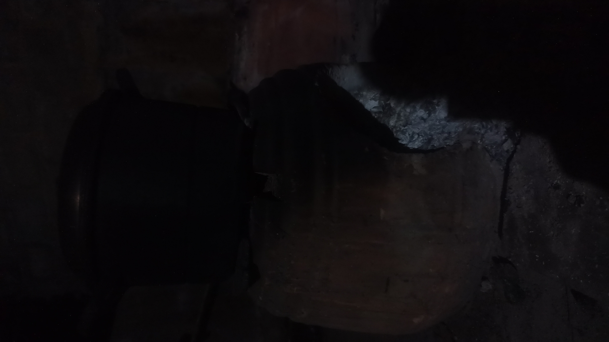 The image shows a dark scene with a large pot placed on top of a broken clay structure. The clay structure appears to be part of a larger vessel or oven, now shattered and revealing rubble and dirt beneath it. The pot is dark and appears to be made of metal, with a rounded lid. A shadow of a person is cast on the right side of the image, partially obscuring the broken clay and the surrounding environment. The overall scene is dimly lit, suggesting it might be nighttime or an indoor setting with limited light. The location is identified as Tuba, Indonesia, though no specific landmarks or contextual details are visible within the image to pinpoint this location further.