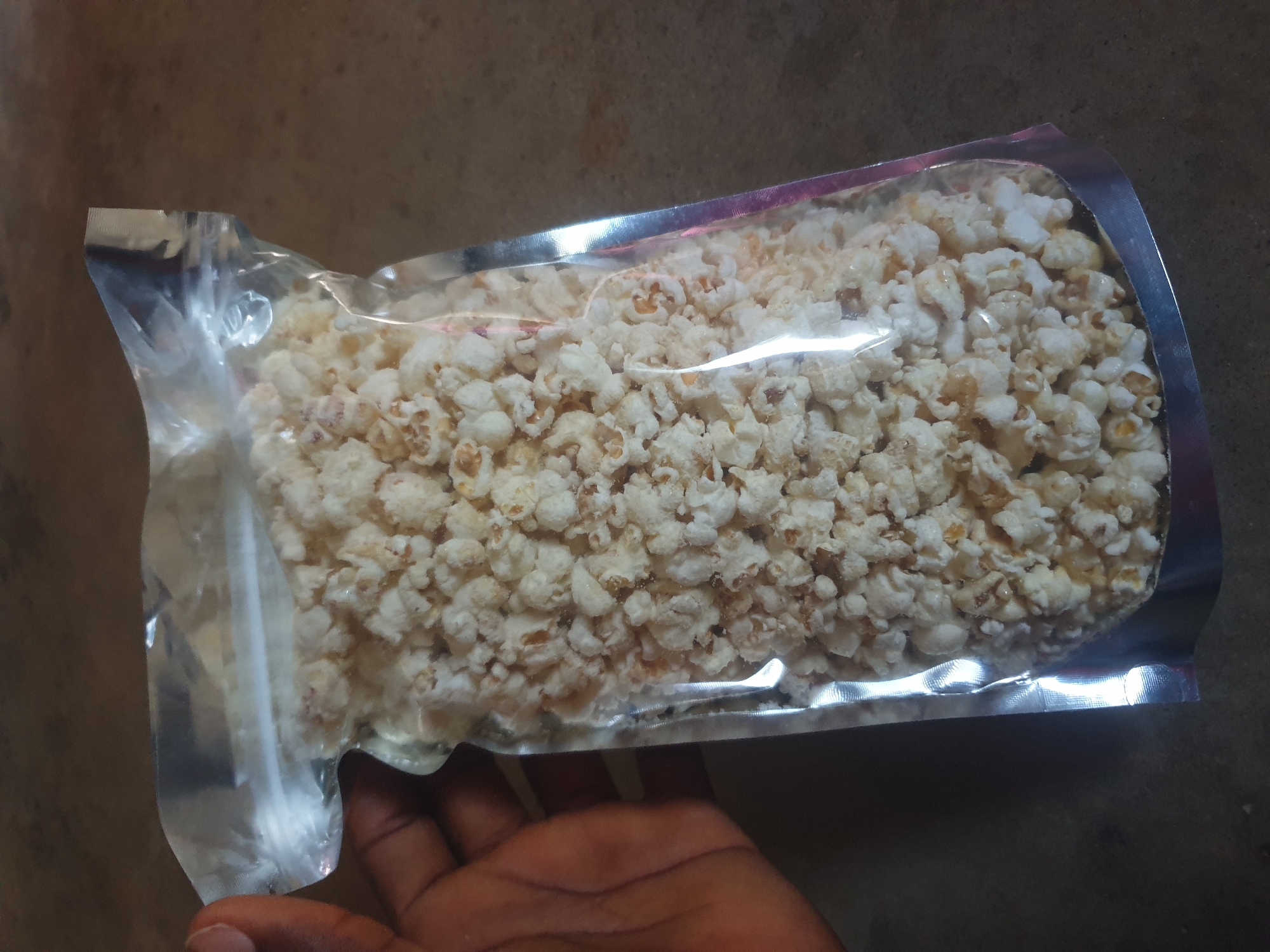 A dark-skinned human hand grasps a transparent, heat-sealed plastic pouch filled with popcorn, positioned slightly off-center to the right. The pouch features a clear upper section revealing the contents, and a metallic, opaque silver-colored base that forms a stand-up bottom. The popcorn inside is primarily white and fluffy, with some kernels exhibiting a faint yellowish or light brown tint. The hand holds the left side of the pouch, with fingers wrapped around the packaging. The background consists of a dark, mottled surface, possibly a floor or tabletop, appearing uniform in texture and color. The scene takes place in the city of Unknown, Unknown.