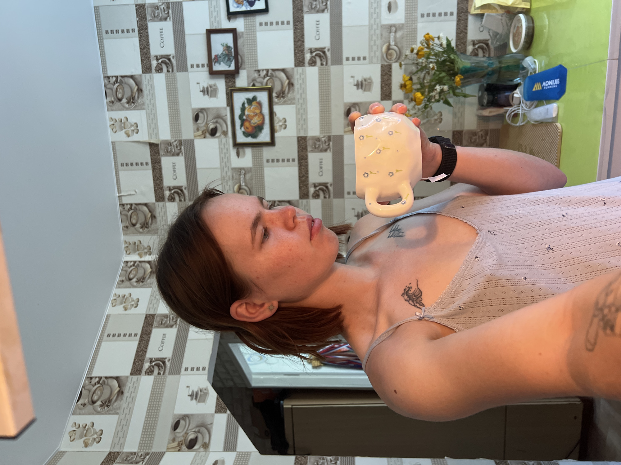 The image depicts a young woman standing in what appears to be a kitchen. She has medium-length brown hair and is wearing a light-colored, thin-strapped top. She is holding a white mug with small blue floral decorations in her right hand, bringing it towards her face as if to drink. A black smartwatch is visible on her left wrist. She has a tattoo of a wave on her chest and another tattoo on her arm.
The background shows a wall with a patterned wallpaper featuring coffee cups and the word "COFFEE" repeated. There are also framed pictures on the wall, one containing oranges and another with a depiction of a figure in a hat. A vase with yellow and white flowers is visible on a green countertop, along with various items, including a blue object with writing on it. To the left, a refrigerator or tall cabinet is partially visible.
The lighting suggests it is daytime, possibly morning, with natural light entering from the right side of the frame. The overall setting is informal and domestic. The provided crops confirm these details, showing the wallpaper patterns, the woman's facial features, her tattoos, and the objects on the countertop more closely. The scene is likely indoors, in a residential kitchen. There is no specific indication of the city of Chaikovsky, Russia within the image itself; this information is provided externally.