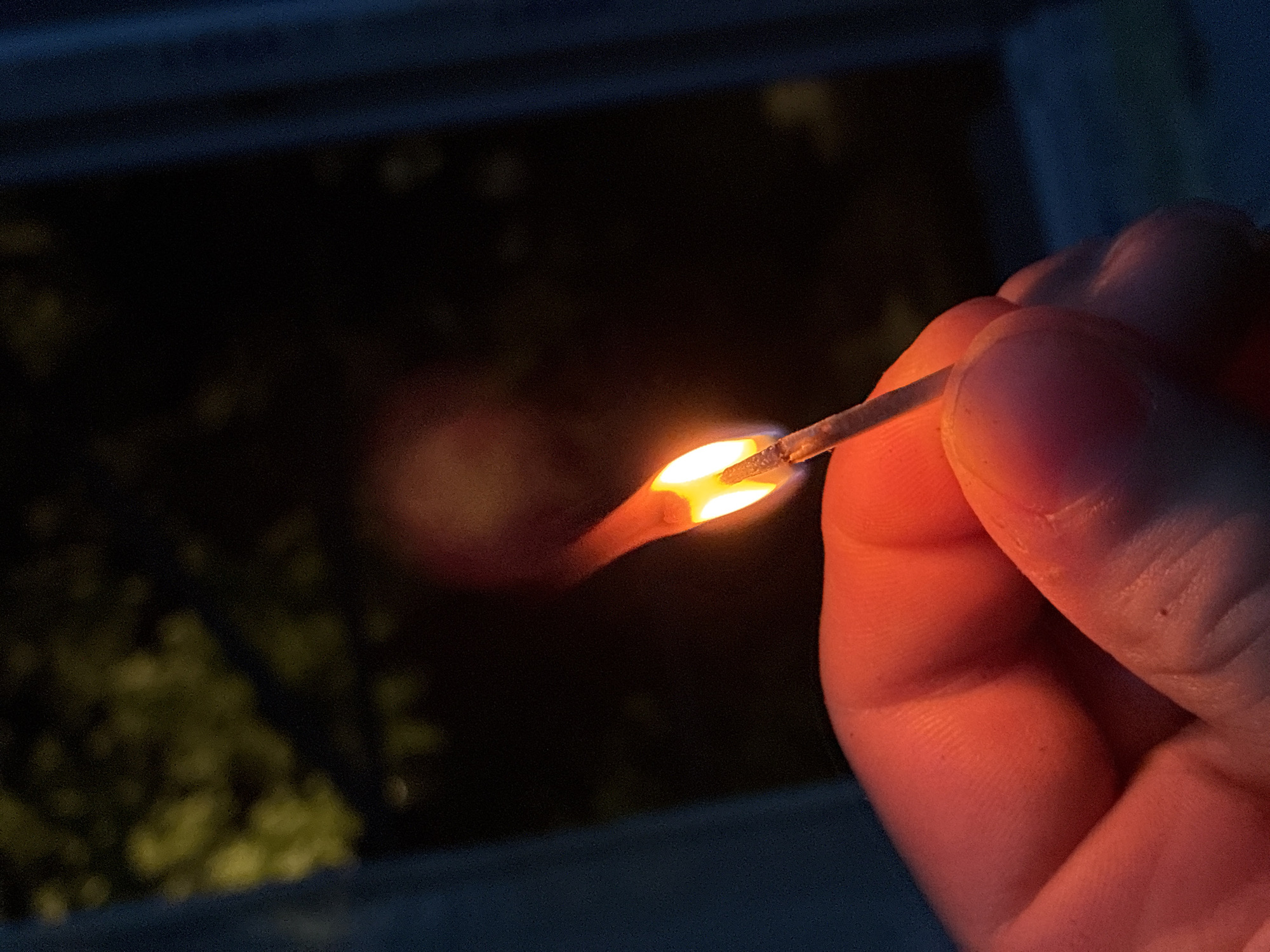 A lit matchstick is held in a hand in a dimly lit environment. The flame of the match is visible and illuminates the fingers holding it. The background is dark, with indistinct shapes suggesting foliage or other objects. The location of Elhovo, Bulgaria, is not discernible from the image.