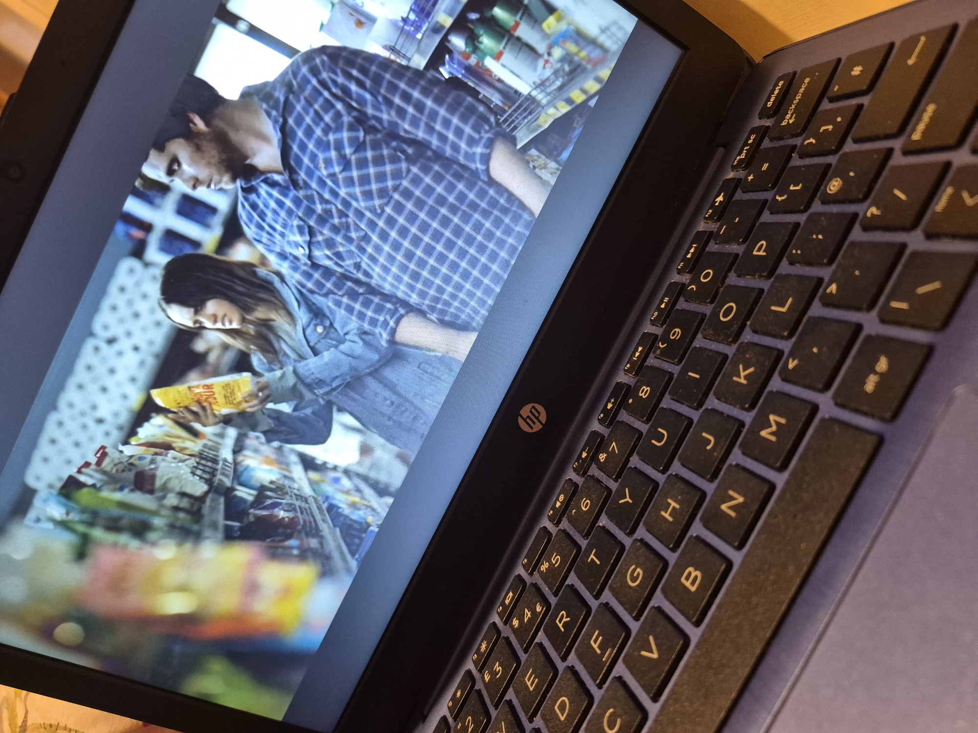 A person is using a laptop, which displays a movie scene. The movie scene shows a woman holding a yellow bag of chips on a shelf in a supermarket, with a man standing next to her. The laptop is on a surface that appears to be a table. The HP logo is visible on the laptop. The image is taken from a high angle, looking down at the laptop and keyboard. The colors in the movie scene are muted, with the woman and man in the foreground and the supermarket shelves in the background. The keyboard has black keys with yellow lettering. The overall lighting is dim, with a warm tone. There is no discernible location in Sandwell, United Kingdom.