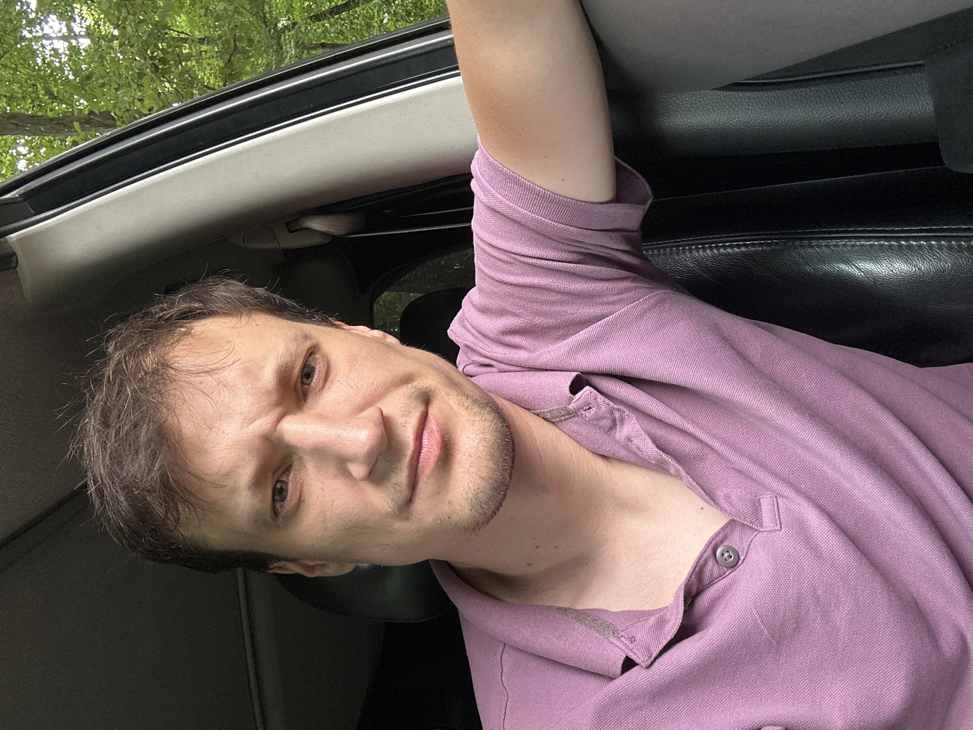 A man with light brown hair and a light complexion is sitting in the driver's seat of a vehicle. He is wearing a light purple polo shirt and has a slight stubble. He is looking directly at the camera with a serious expression, his eyebrows furrowed. The interior of the car is visible, including a black leather seat and the car's door frame. Outside the car window, green trees are visible, suggesting a location in a wooded area, possibly near Shatava, Ukraine. The image is a close-up selfie taken from the man's perspective.