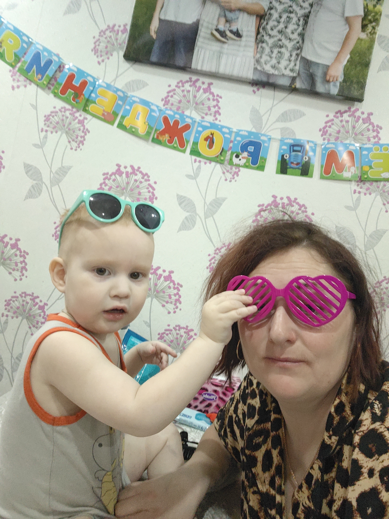 The image shows a woman and a young child indoors, likely at a birthday celebration. The woman is wearing pink heart-shaped sunglasses and a leopard print shirt. The child is wearing teal sunglasses on their head and a tank top. The child is reaching towards the woman's face. A "Happy Birthday" banner in Russian hangs on the wall behind them, above a framed family photograph. The location is in Markovskii, Russia, based on the Russian text in the banner.