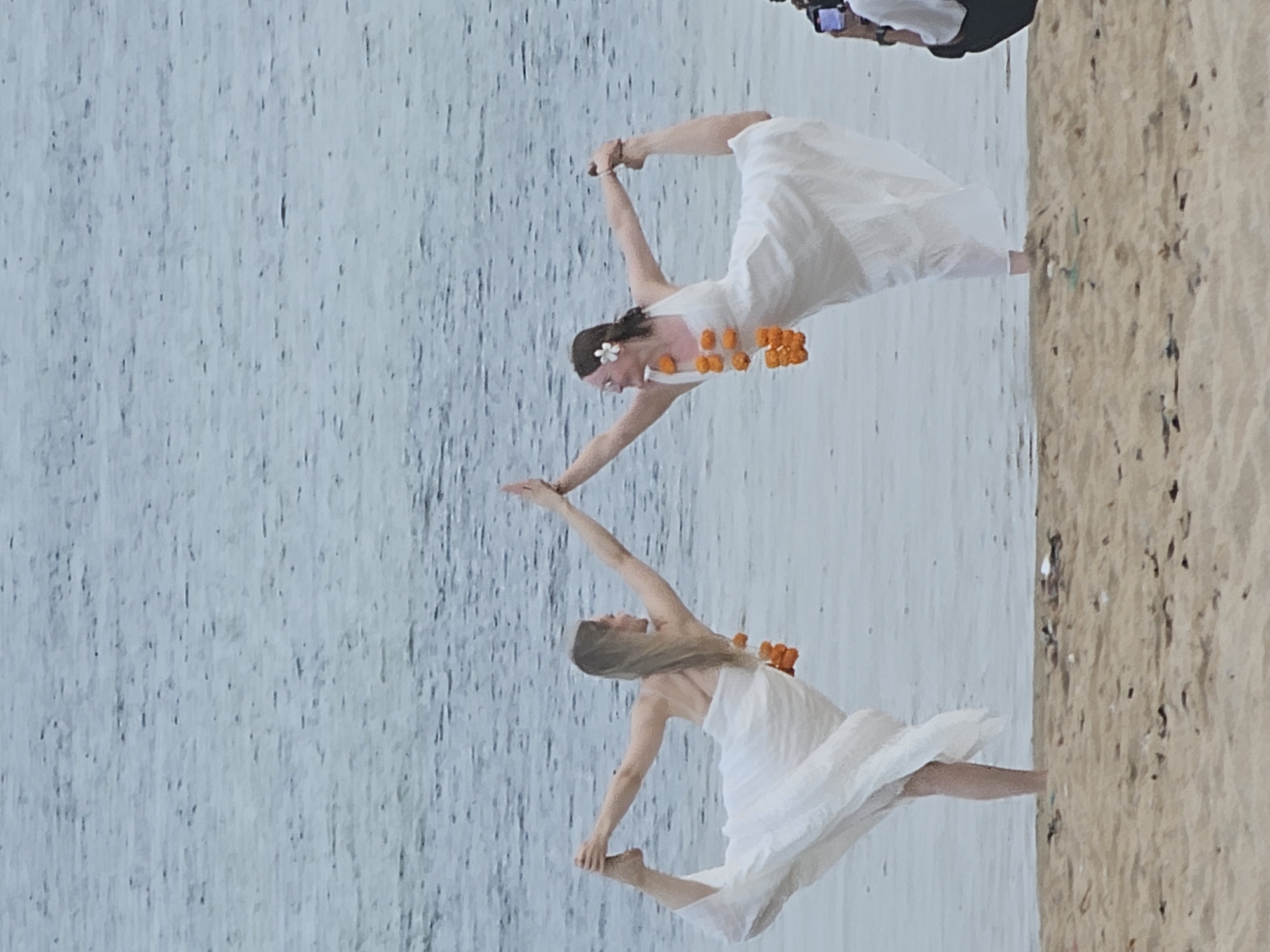 Two women in white dresses are practicing yoga on a sandy beach, with a body of water in the background. The woman on the left has her left leg extended behind her, with her left foot held by her left hand, while her right arm is extended upwards. The woman on the right is mirroring this pose, with her right leg extended behind her, her right foot held by her right hand, and her left arm extended upwards. Both women are adorned with garlands of orange flowers around their necks and the woman on the right has a white flower in her hair. The beach appears to be littered with debris. In the background, a person is partially visible on the right, holding a camera. The scene is set at Sanur Beach in Denpasar, Indonesia.