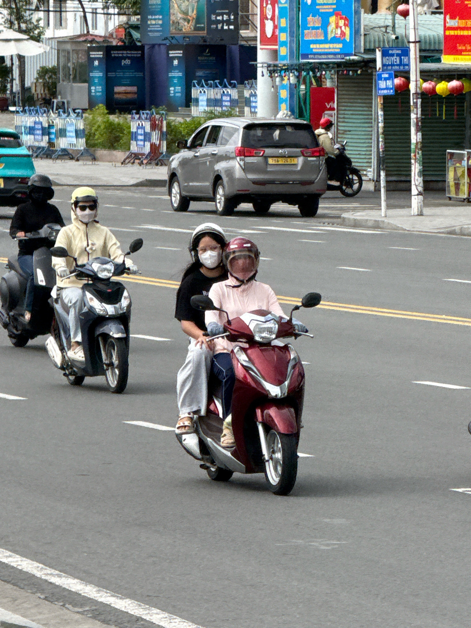 The image depicts a street scene in Nha Trang, Vietnam, likely on a clear day. Multiple motorbikes are visible, a common mode of transportation in the city.
In the foreground, two women are riding on a dark red scooter. The woman in front, wearing a light pink jacket and a helmet with a visor, is driving. The woman behind her, wearing a black shirt and a helmet, is a passenger. Both are wearing face masks.
Behind them, a person in a black jacket and helmet is riding a dark grey scooter. Further back, a person in a light yellow hooded jacket, sunglasses, and a face mask is riding a dark blue scooter. This scooter also has a black front and silver accents. The rider is wearing white gloves.
In the background, a silver SUV is parked on the right side of the road, with a license plate that reads "79A-126.01". Across the street, there are buildings with signs, including what appear to be advertisements and street signs with Vietnamese text. Several blue and white striped barriers are also present on the sidewalk. The road itself is paved and has white lane markings. Red and yellow lanterns are hanging from a pole on the right side of the image.