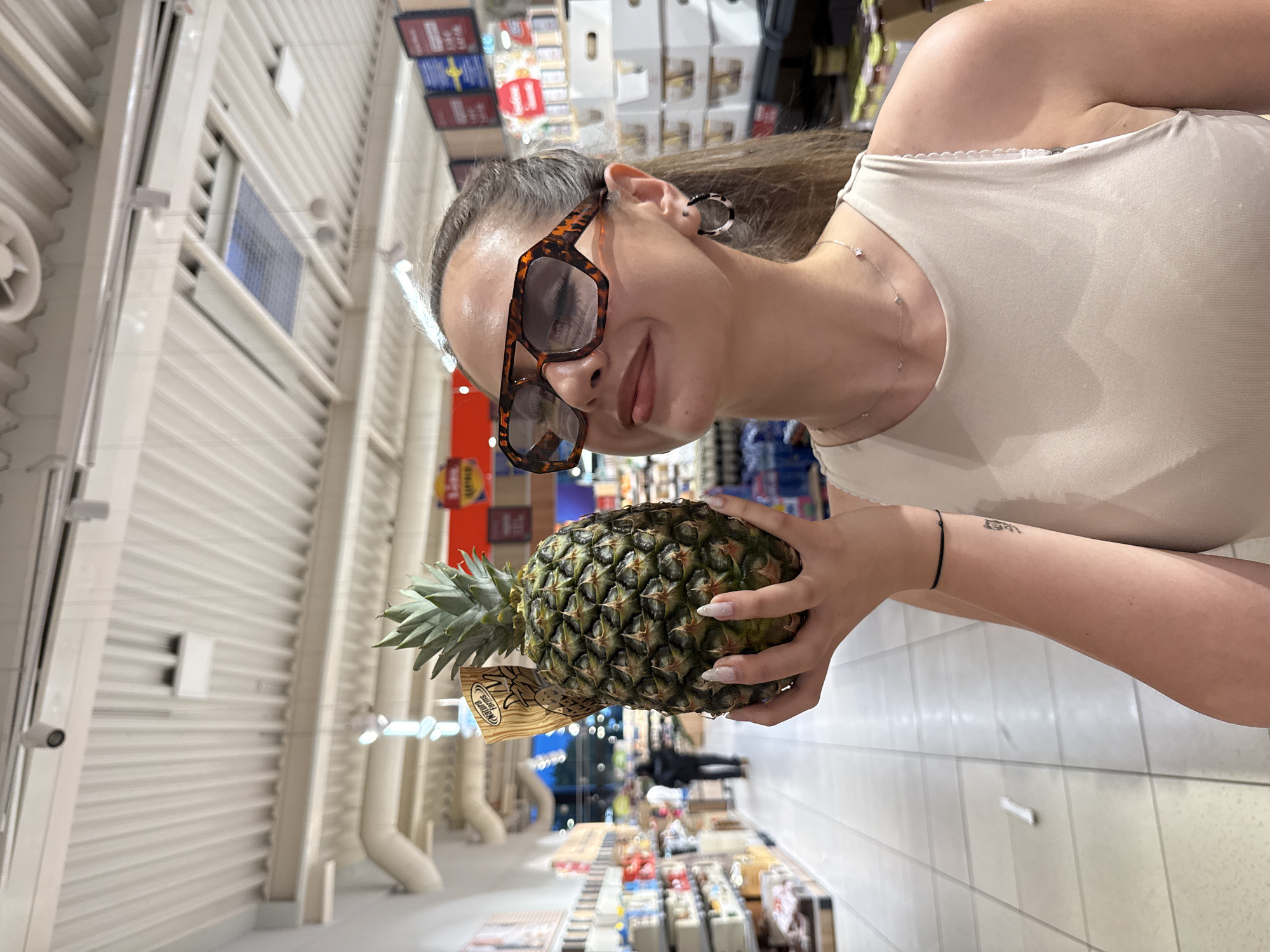 A young woman stands in a supermarket aisle, holding a pineapple. The aisle is lined with shelves of various products. The woman wears sunglasses and a light-colored top. Her arm is visible, adorned with a bracelet. The supermarket is located in Blagoevgrad, Bulgaria.