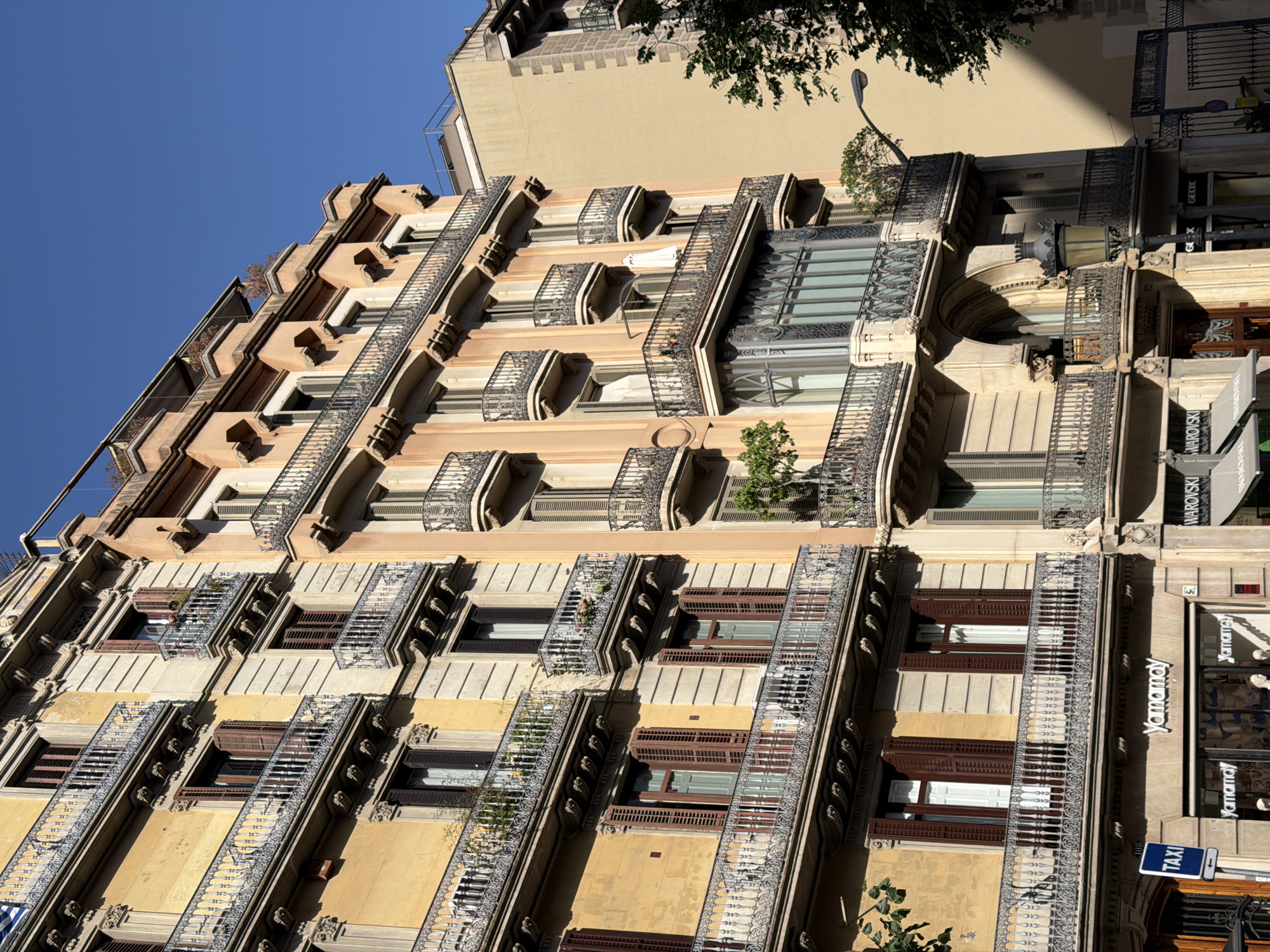 This is a tilted, eye-level, medium shot of a building's facade, likely in Barcelona, Spain, based on the provided context. The building is made of light-colored stone with several floors, each featuring a series of balconies with ornate metal railings. The balconies are adorned with plants, adding a touch of greenery to the architecture. The windows have dark wooden shutters, some of which are open, revealing glimpses of interiors. The facade is a mix of light yellow and cream tones, with strong shadows indicating it's a sunny day, likely late morning or early afternoon.
At street level, there are shopfronts. Visible text includes "Yamamay" appearing multiple times, "SWAROVSKI," and "GEOX," suggesting these are retail stores. A blue sign with "TAXI" is also visible. The overall impression is of a well-maintained, possibly historic, urban building in a commercial district. The clear blue sky at the top of the frame and the shadows suggest pleasant weather.