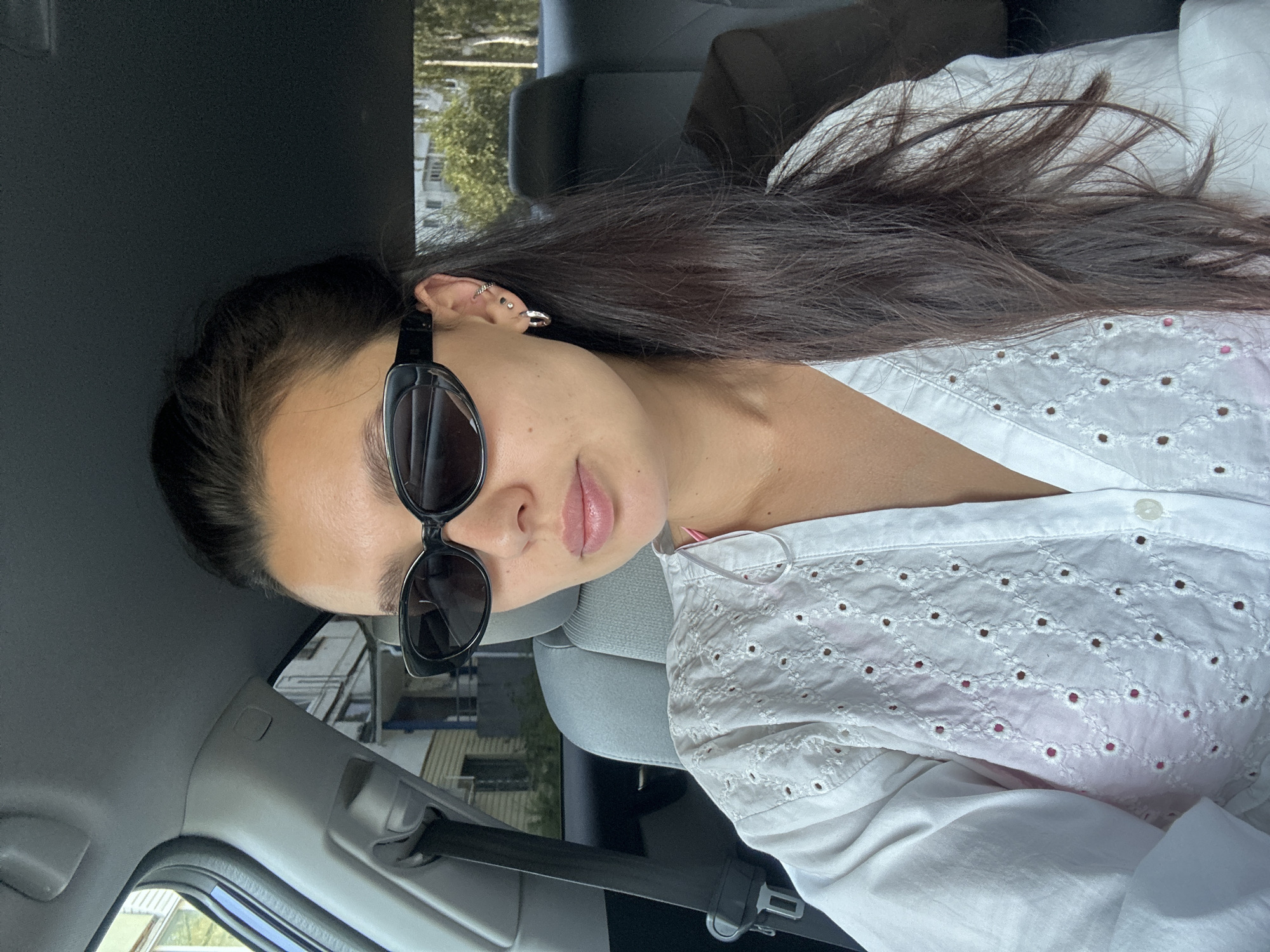 A young woman with dark hair pulled back into a ponytail is sitting in the passenger seat of a car. She is wearing black sunglasses and a white broderie anglaise top over a pink garment. She has small hoop earrings and a silver stud in her left ear. The car interior is visible, including the grey seat, seatbelt, and the car's ceiling. Outside the car windows, buildings and trees suggest an urban or suburban environment. The image appears to have been taken during daylight hours. The specific location of Chaikovsky, Russia, cannot be determined from the visual information provided in the image.