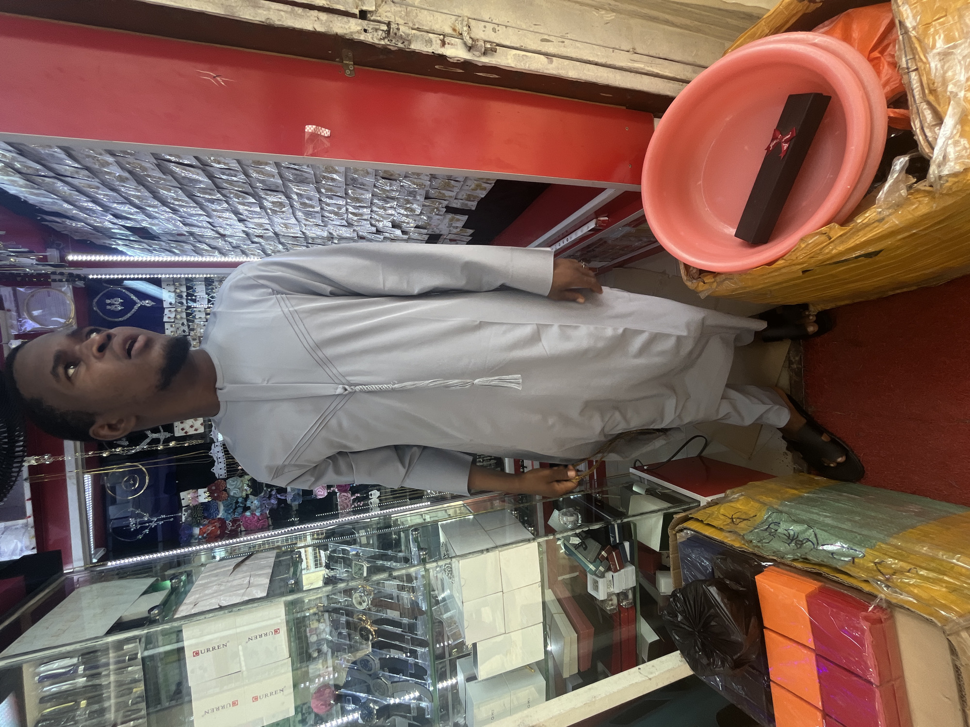 In a dimly lit shop, a young Black man stands looking to his right, wearing a light grey traditional long-sleeved tunic with intricate stitching on the chest and a decorative tassel. He is barefoot in dark sandals. The shop is filled with display cases and shelves stocked with various items, including watches, jewelry, and what appear to be packaged accessories. To his left, a glass display case shows rows of watches. To his right, a red wall is covered in small, individually packaged items, possibly jewelry or accessories. In the foreground, to the man's left, are several stacked boxes, some wrapped in plastic, containing colorful gift boxes. To his right, a large pink plastic basin holds a dark rectangular box with a red ribbon. The overall impression is of a retail environment, likely a small jewelry or accessory shop in Abuja, Nigeria.