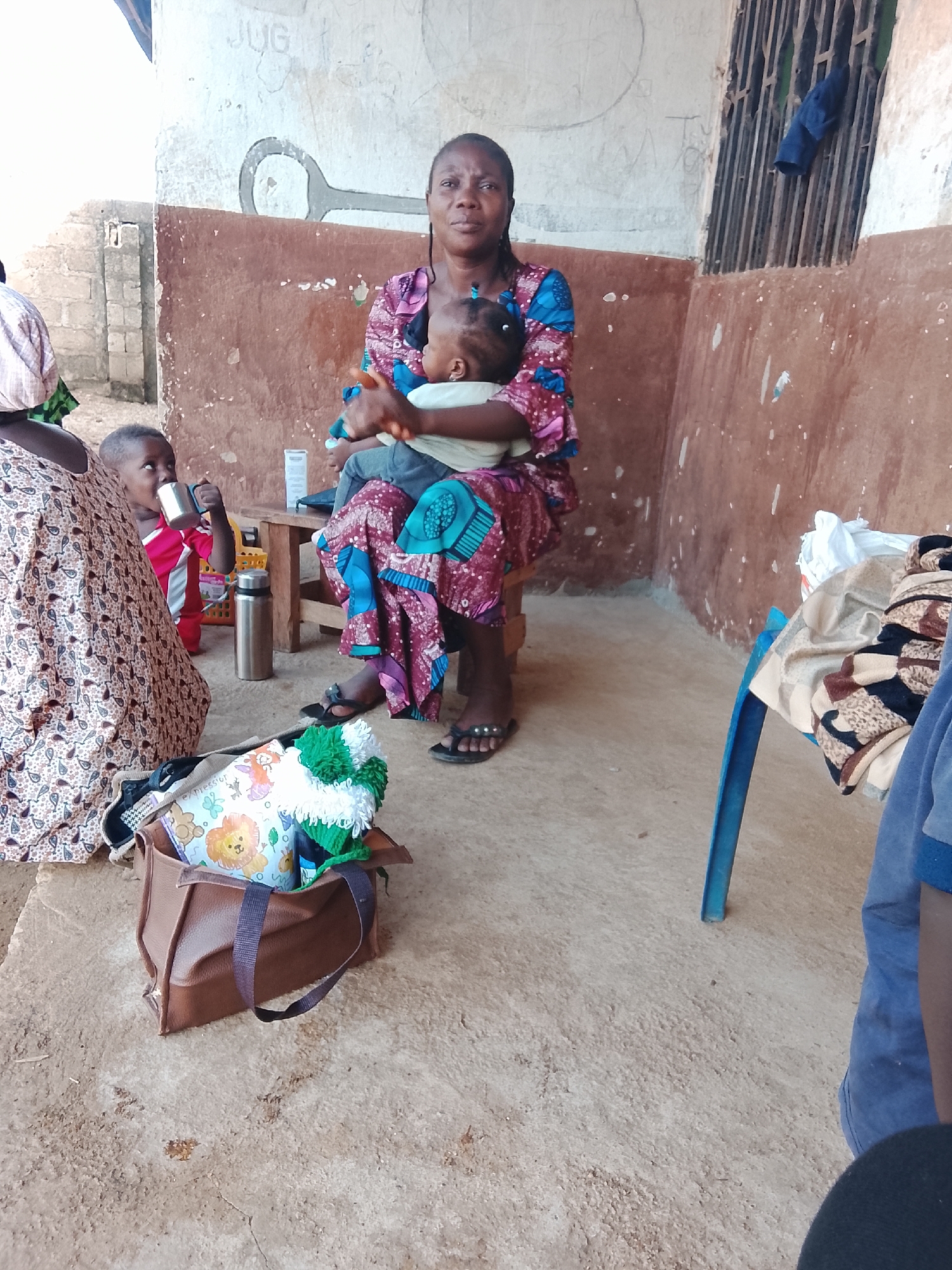 The image depicts a scene outdoors, likely in Akwanga, Nigeria. A woman is seated, holding a baby. Beside her, a young child drinks from a metal cup. In the foreground, a brown bag lies on the ground, containing fabric items. A blue chair is visible to the right, draped with various textiles. The background shows a textured wall with some writing and a barred window. The ground is a rough, sandy surface.