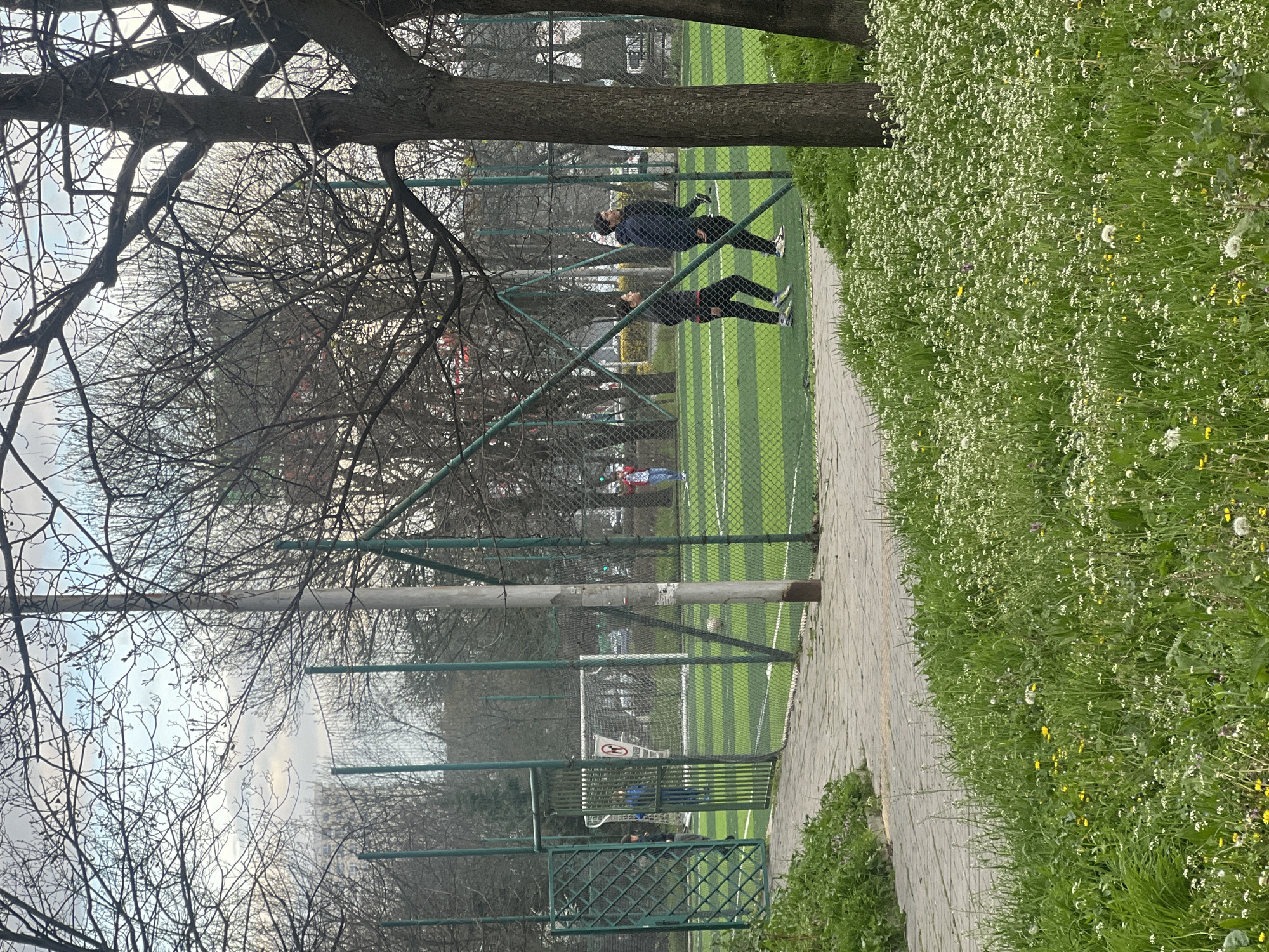 The image depicts a partially obstructed view of a soccer field in Burgas, Bulgaria, seen through a fence and tree branches. On the field, two people are visible near the fence. A paved walkway runs parallel to the fence, with a field of wildflowers to the right. The background features more trees and what appears to be a cloudy sky. A soccer goal is visible on the field.