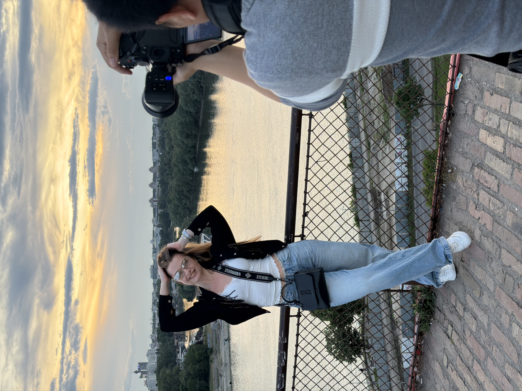 A woman in jeans, a white top, and a black jacket lies on a brick and metal surface, smiling at the camera. She has her hands behind her head. An individual holding a camera is visible above her. The background shows a river and cityscape under a bright sky. The location is Belgrade, Serbia, as suggested by the skyline and architecture.