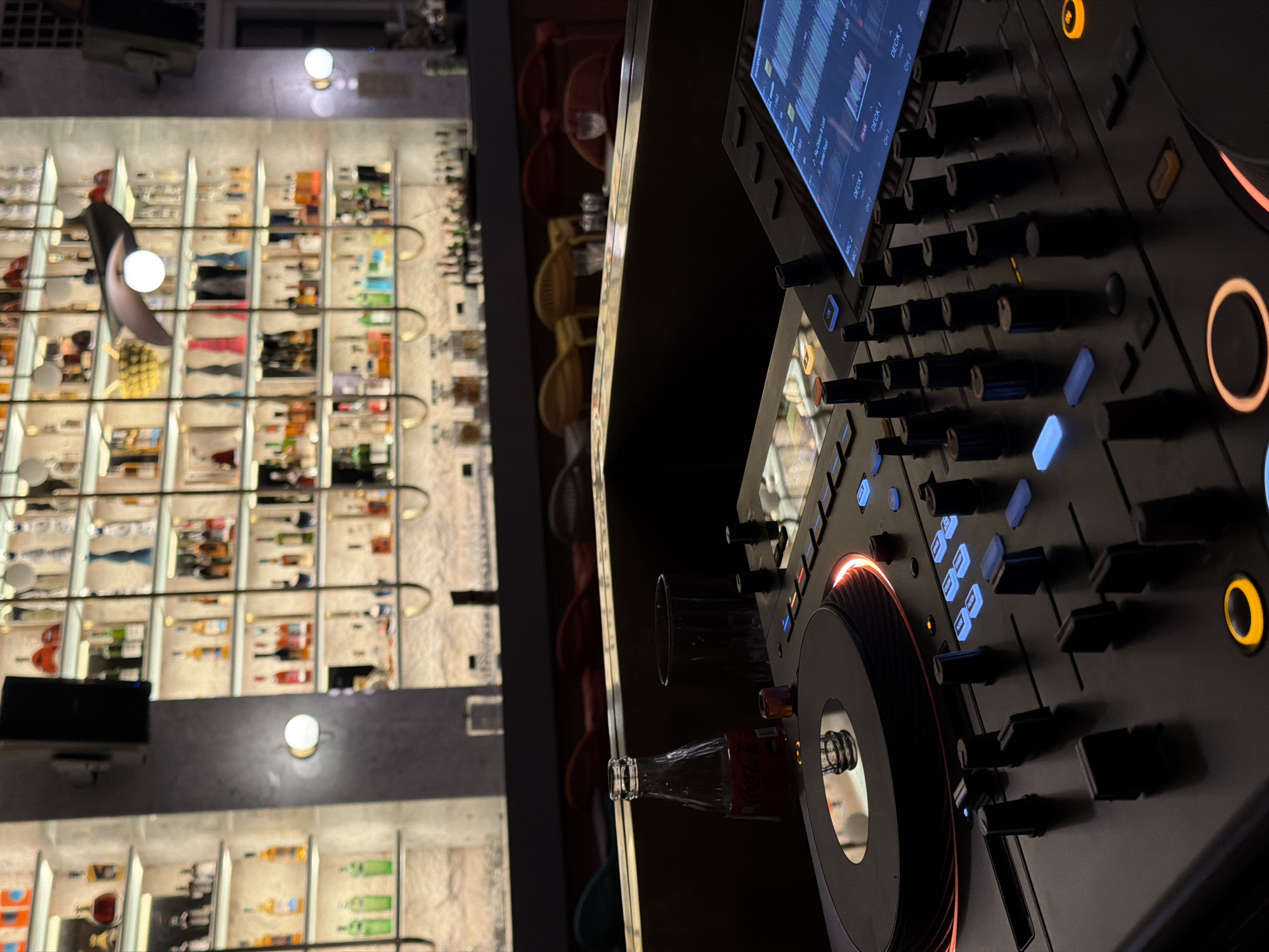 The image shows a DJ setup in a bar, likely in Budva, Montenegro, based on contextual inference. The setup includes a modern DJ controller with a screen displaying waveforms and various controls, such as knobs, faders, and illuminated buttons. A glass of liquid and a glass bottle of Coca-Cola are adjacent to the controller. The backdrop features a wall-mounted liquor display with illuminated shelving showcasing numerous bottles of alcohol. The lighting in the bar is a combination of ambient lighting from the display and the illuminated elements of the DJ equipment.