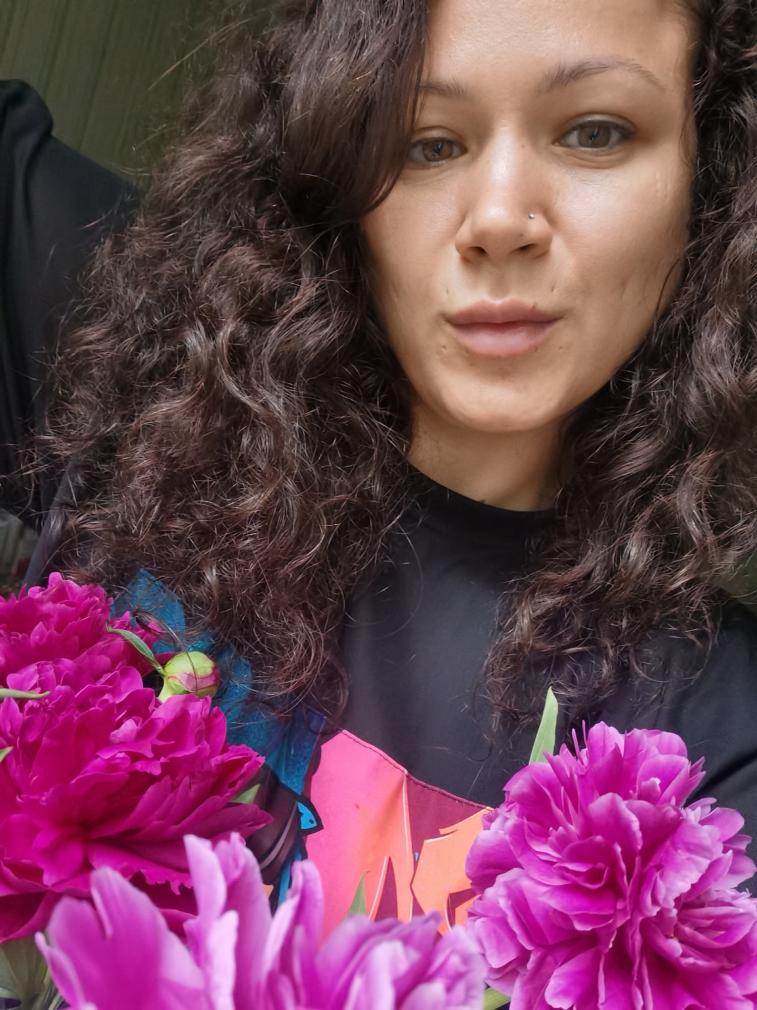A close-up selfie of a young woman with dark curly hair and a nose piercing is shown. She is wearing a black t-shirt. In the foreground, there are several vibrant pink peonies. The background is blurred and appears to be indoors, with hints of a wall and possibly fabric. The image was taken in Chaikovsky, Russia.