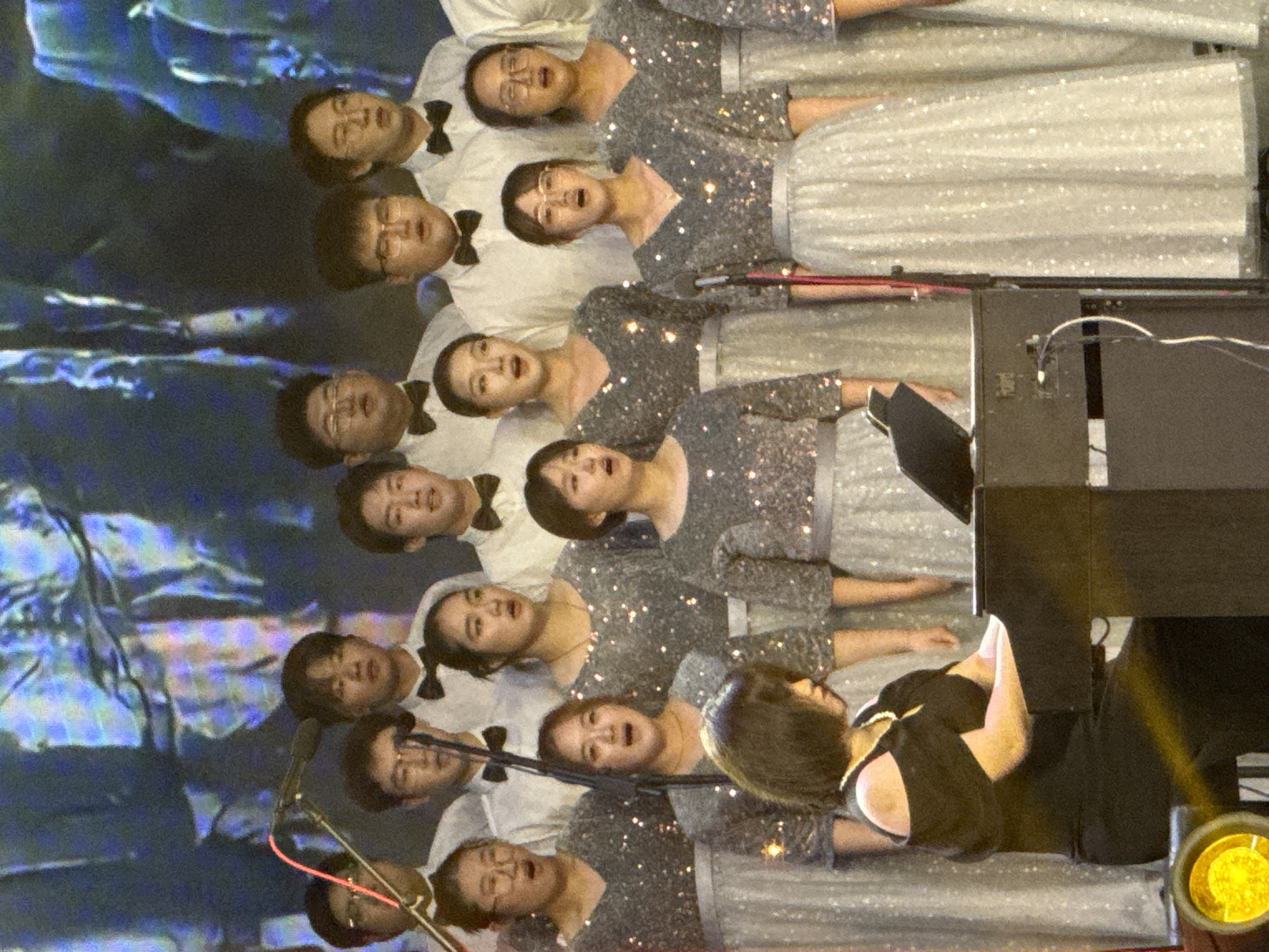 An indoor concert scene shows a choir performing on stage in Guta, China. The image, rotated 90 degrees clockwise, features multiple rows of choir members, predominantly facing towards the right side of the frame. The women in the front rows wear matching sparkly grey tops with light-colored, shimmering long skirts, while the men in the back rows are dressed in white shirts with black bow ties. Many of the choir members have their mouths open, indicating they are singing.
In the foreground, to the right of the choir, a person, likely the conductor or a pianist, is seen from the back, wearing a black top and black trousers. A black digital piano or keyboard is positioned next to this individual. Several microphones on stands are visible among the choir members. The background is a large screen displaying abstract blue and purple light patterns, suggesting a dynamic stage setting. A bright yellow spotlight illuminates the scene from the far right. The overall atmosphere appears to be a formal musical performance. No discernible text is visible in the image.