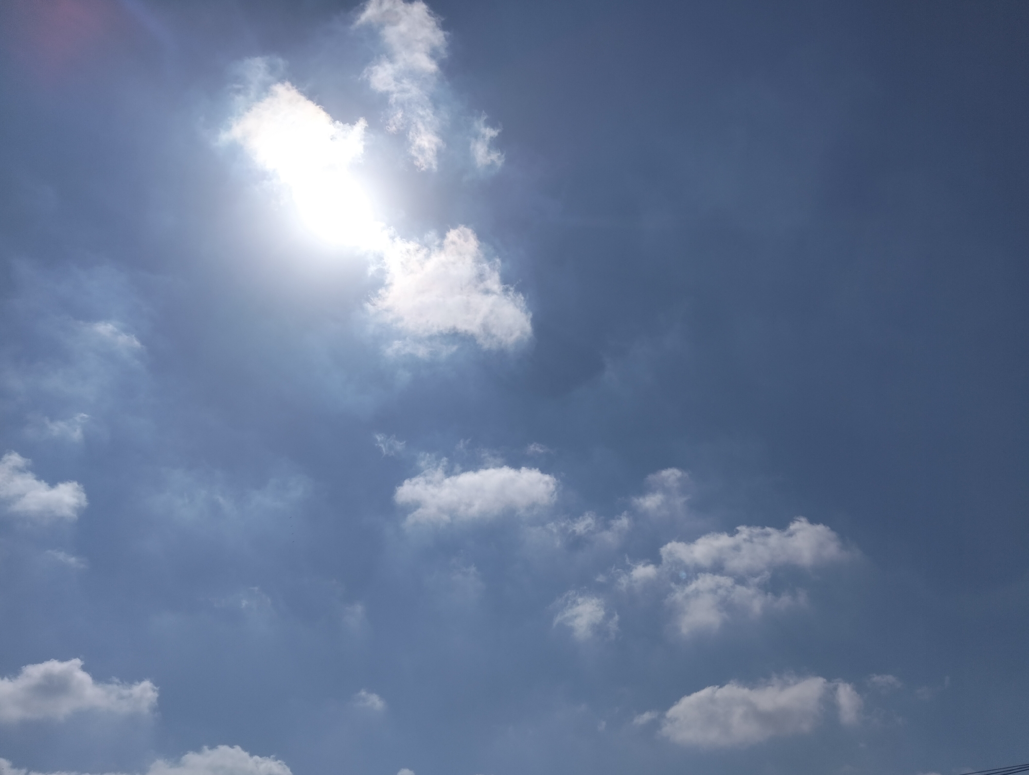 The image captures a bright, sunny day with a clear blue sky in Kiryat Ata, Israel. The sun is prominently visible in the upper left quadrant, casting a strong glare and its rays are partially obscured by scattered white clouds. Several clouds of varying sizes and densities are distributed across the sky, some appearing fluffy and white, while others are darker and more diffuse, suggesting varying levels of moisture. There are no people, man-made structures, or discernible ground features visible in this outdoor shot. The scene appears to be an unpopulated sky, likely taken from an open area within the city.