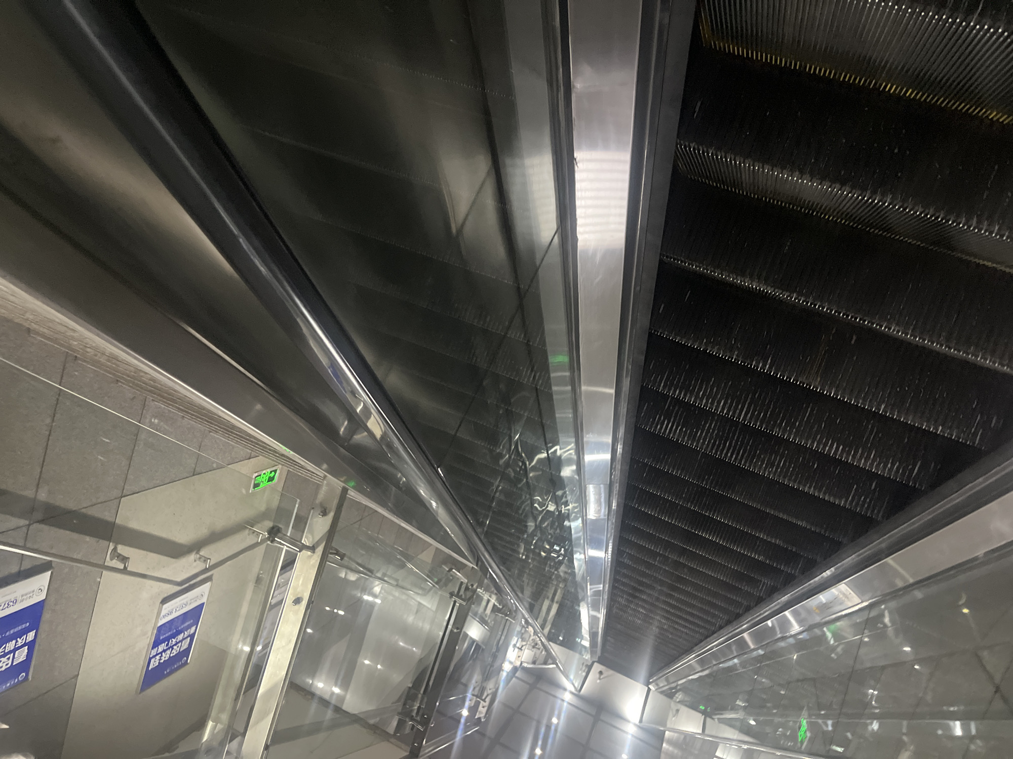 An upward-angled, wide shot shows two parallel escalators, one on the left and one on the right, within what appears to be a modern subway station in Yuzhong District, China. The escalators are made of dark gray metal steps with silver metal sides and handrails. Both escalators are illuminated by overhead fluorescent lights, and there are green illuminated signs with arrows on the left escalator's glass barrier. The walls of the station are a light gray concrete, and there are framed posters with Chinese text on the right wall. One poster features a blue background with white Chinese characters and a telephone number. The overall environment is clean and well-lit, suggesting a public transportation hub. There are no people visible in the image.