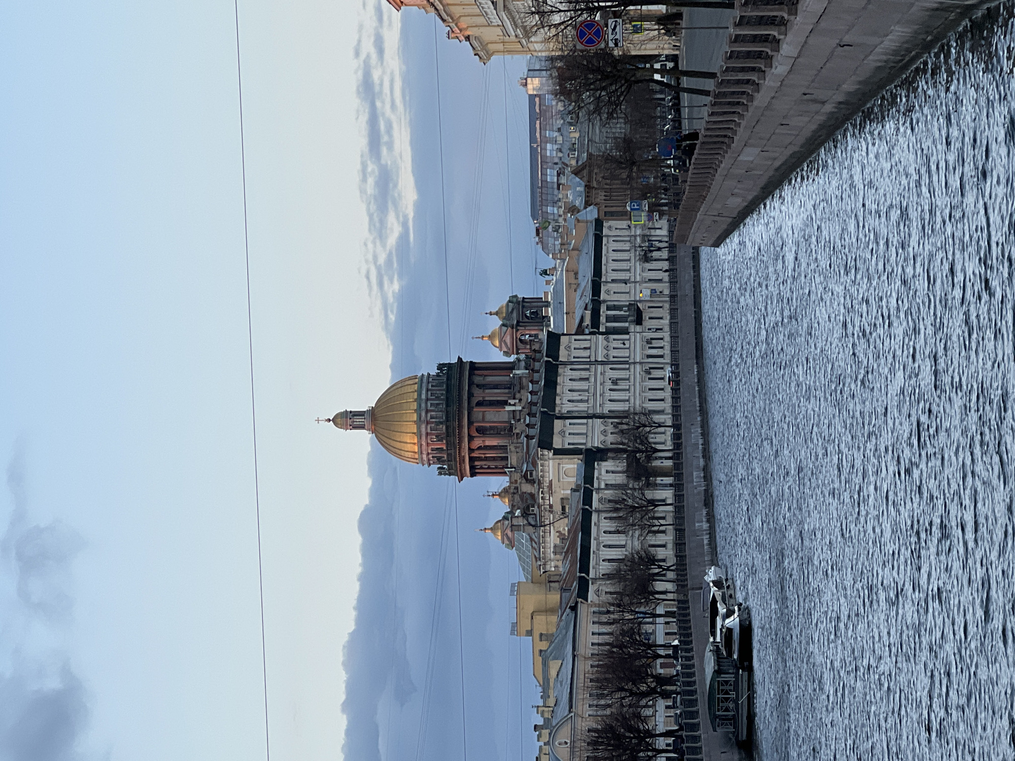 The image presents an urban landscape scene in Saint Petersburg, Russia, likely captured during late afternoon or on an overcast day. The view looks across a wide canal or river, with the city's architectural landmarks lining its banks.
The most prominent subject is a grand, classical cathedral-like structure, positioned on the left side of the frame. It features a large, luminous golden dome along with several smaller domes, towering above other buildings. The architecture is detailed, with columns and multiple tiers.
To the right of the buildings, a wide canal or river dominates a significant portion of the frame, with its surface showing ripples in dark blue-grey tones. Along the near bank of this waterway, there is a paved embankment or street. This bank is lined with several multi-story buildings, characterized by their classical facades and numerous windows. Bare trees are sparsely visible along the embankment, suggesting winter or early spring. Several white boats are docked along the embankment.
The sky above is mostly overcast or partly cloudy, displaying muted tones of light blue and grey. Thin power lines are visible against the sky. No specific human activity is discernible, giving the scene a calm, still atmosphere.
Notable details include a few traffic signs on the embankment; one clearly visible sign is a red circle with a blue cross, indicating "No Stopping" or "No Standing," and another is a blue square with a white "P," signifying "Parking."