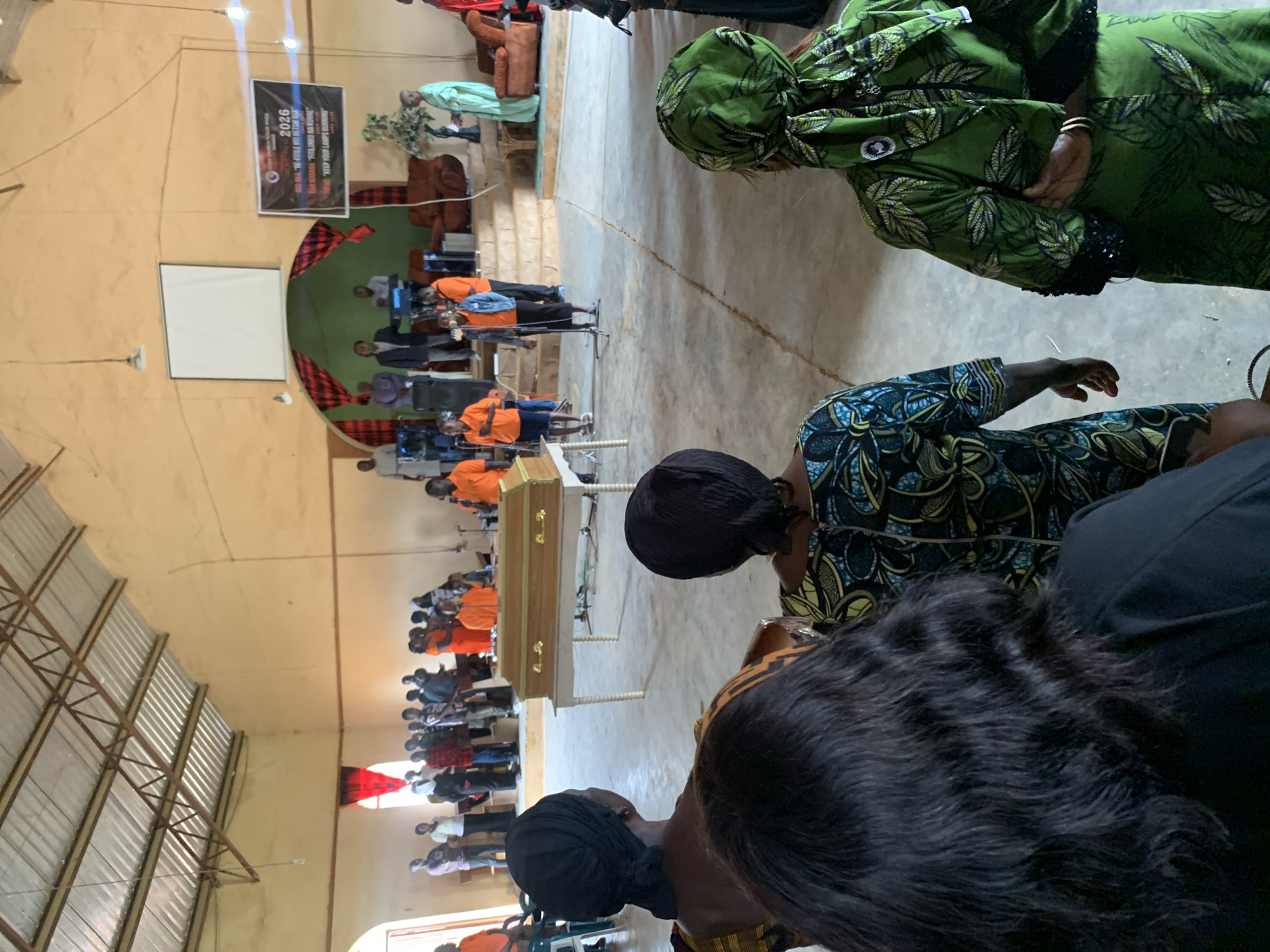 In Miango, Nigeria, a funeral service takes place inside a church with a metal roof and cream-colored walls. At the front of the church, a light brown wooden casket rests on a white table. Several people stand behind the casket, some in bright orange shirts, while a man in a suit stands at a podium. A large banner hangs on the wall behind them, displaying "2026" prominently. In the foreground, three women, facing away from the camera, are seated, observing the service. Two of the women are wearing headscarves, and all three are dressed in traditional African attire. The church floor is concrete, with a visible crack running across it.