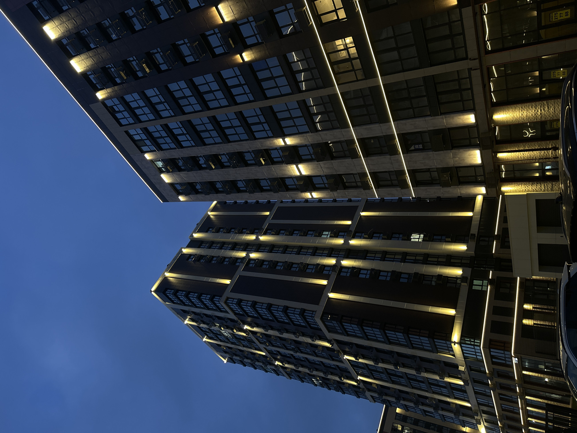 The image displays a nighttime view of two illuminated modern high-rise buildings in Rostov-on-Don, Russia, under a dark blue sky. The building on the left is taller, featuring a dark facade with vertical light-colored panels and numerous dark-paned windows. It is accented with vertical warm-toned strip lighting along its edges and recesses. The building on the right presents a multi-toned facade of dark and lighter gray/beige panels, also with dark windows and balconies. This building is illuminated by both horizontal strip lighting and vertical wall-mounted fixtures. At the ground level, commercial spaces are visible with large windows and additional lighting. The tops of several dark-colored vehicles are partially visible at the bottom of the frame. A white sign with "АРЕНДА" (Rent) and a phone number (275 77 88) is visible on the lower right building, alongside a glowing stylized symbol resembling a bull's head on a brick-textured wall.