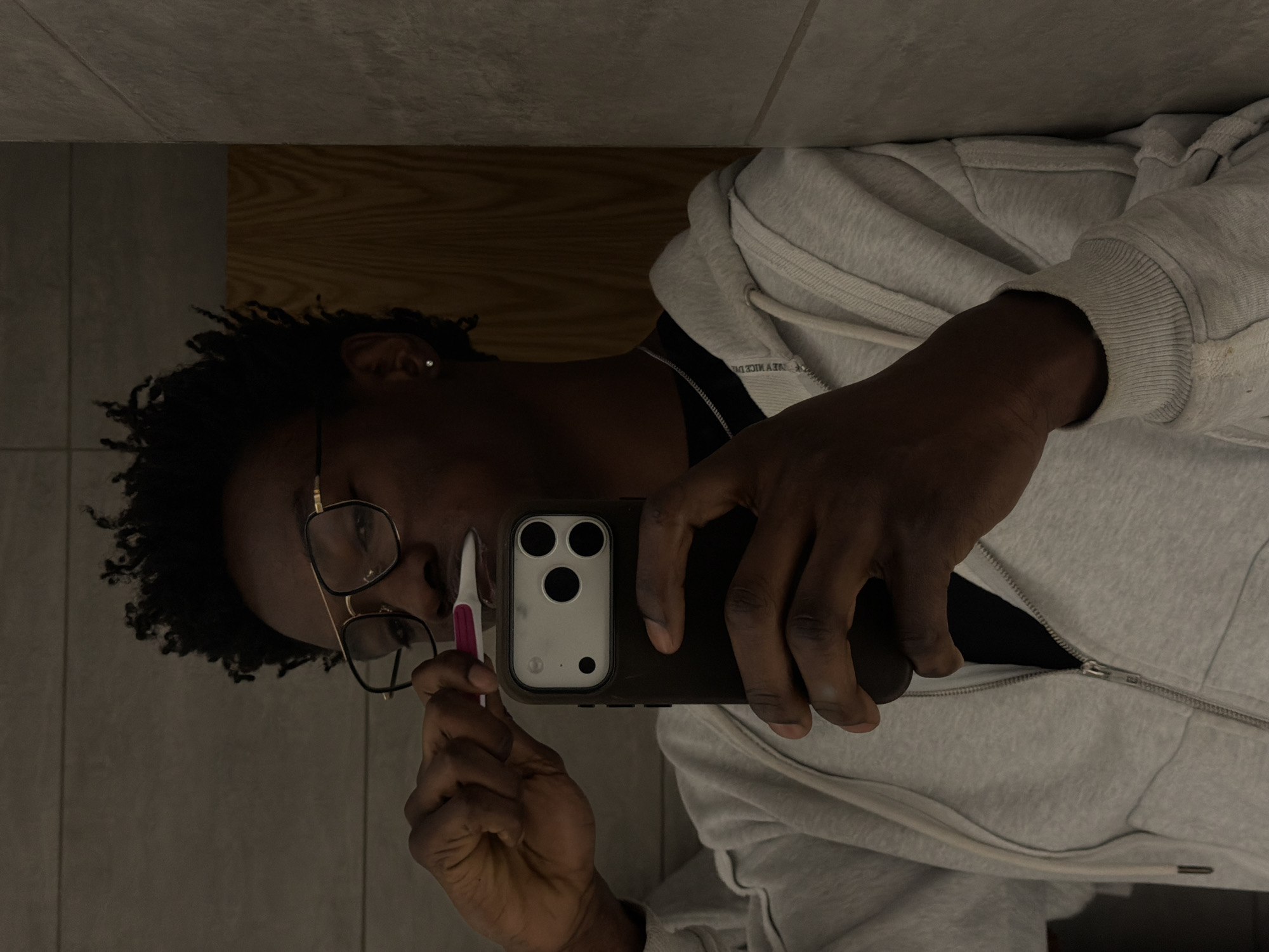 The image displays a mirror selfie of a person in an indoor setting, likely a bathroom in London, United Kingdom. The individual has dark skin, dark curly hair, and wears gold-rimmed rectangular glasses. They are dressed in a light gray zipped hooded sweatshirt, with a black undershirt visible beneath. A small, silver stud earring can be seen in their right ear.
The person is holding a black smartphone with a triple camera setup, which is being used to capture the reflection. In their other hand, they hold a small white and pink object, resembling a toothbrush or a facial tool, near their face. Their expression is neutral and focused on the task at hand.
The background consists of light gray tiled walls on the left and a wooden surface, possibly a vanity or counter, on the right. The lighting appears dim. Faint, inverted text is visible on the light gray hoodie, but it is largely illegible.