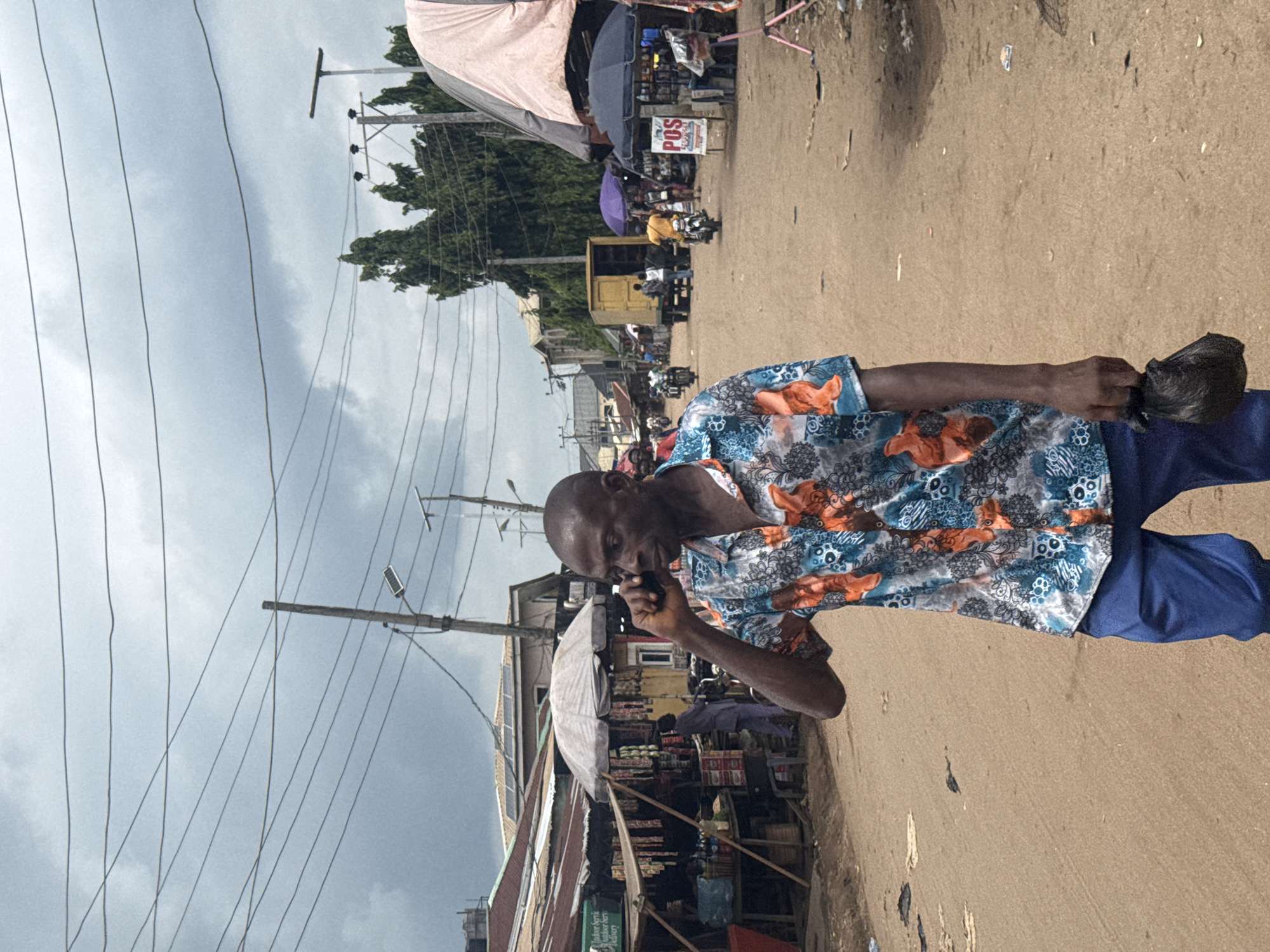 The image shows a man in Ihiagwa, Nigeria, holding a dark object, possibly a bag, and talking on a cell phone. He is wearing a patterned shirt with blue pants. The scene appears to be a busy street with shops and vendors. The background shows utility poles and wires, as well as various buildings and structures. The sky is overcast. The ground is a reddish-brown color.