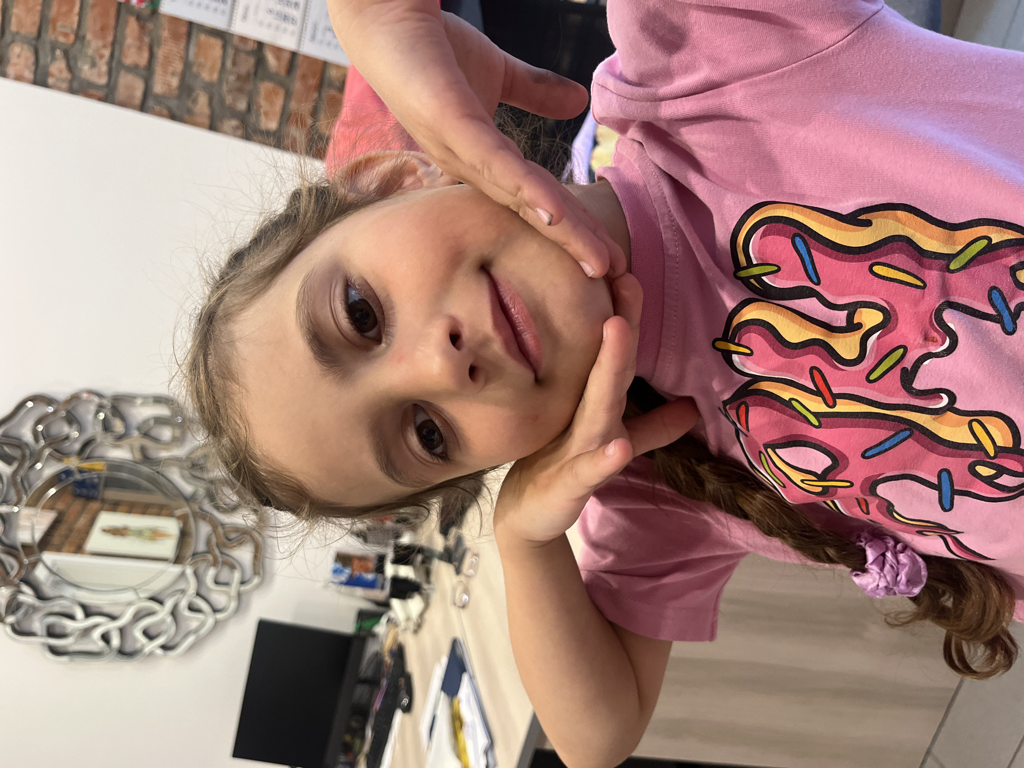 The image shows a close-up, over-the-shoulder shot of a young girl with brown hair in a braid, wearing a pink t-shirt with a colorful donut graphic. She is resting her chin on her hands and looking at the camera with a slight smile. The background is blurred, but suggests an indoor setting with a desk, computer monitor, and brick wall. It is impossible to determine the location within Svetlogorsk, Russia.