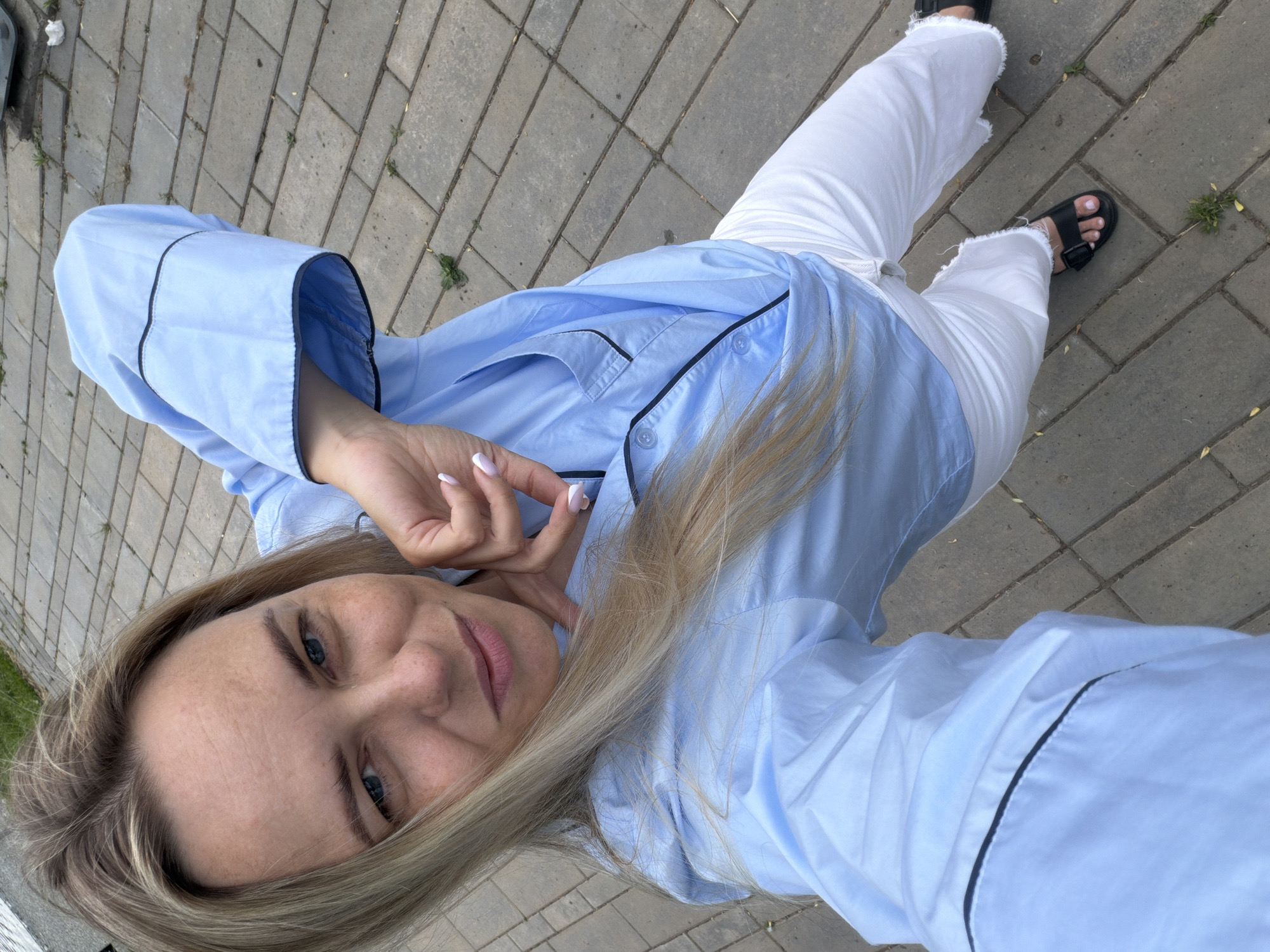 A high-angle selfie captures a blonde woman outdoors in Votkinsk, Russia. She looks directly at the camera with a gentle, relaxed smile. Her long, light blonde hair is parted to the side, framing her face, which has blue eyes and a light complexion with some visible freckles. She wears a light blue, pajama-style button-down shirt featuring dark black piping along the collar, placket, and sleeve cuffs. Her nails are manicured with a light, possibly French-tip style, polish. One hand is gently resting near her chin.
She is also wearing white, relaxed-fit pants with distressed, frayed hems, paired with black sandals that have a buckle detail. The setting is an outdoor paved area, likely a sidewalk or pedestrian path, composed of grey rectangular concrete blocks. Small patches of green vegetation are visible growing in the crevices between the blocks. The lighting suggests it is daytime, appearing soft with no harsh shadows, indicating possibly an overcast day or shaded area. There is no visible text in the image.