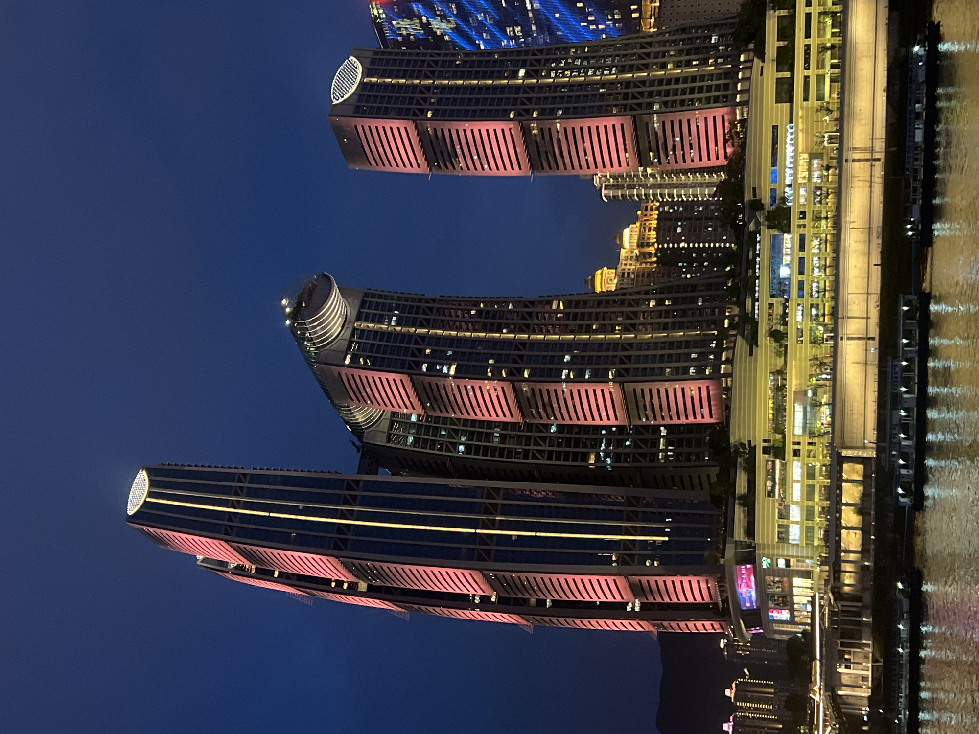 The image shows a waterfront view of modern skyscrapers at night in the Jiangbeicheng Subdistrict, Chongqing, China. The buildings are illuminated with vibrant pink and yellow lights, highlighting their contemporary architectural designs. The tallest skyscraper, on the left, has a distinctive curved top and vertical lighting. To its right, a cluster of interconnected buildings features similar angled designs and illuminated facades. Further to the right, another prominent skyscraper with a rounded top is visible, also bathed in light. In the foreground, a wide body of water reflects the city lights. Several boats are docked or moving on the water. The background shows more buildings and a dark, cloudless sky. The scene suggests a lively urban environment at dusk or night.