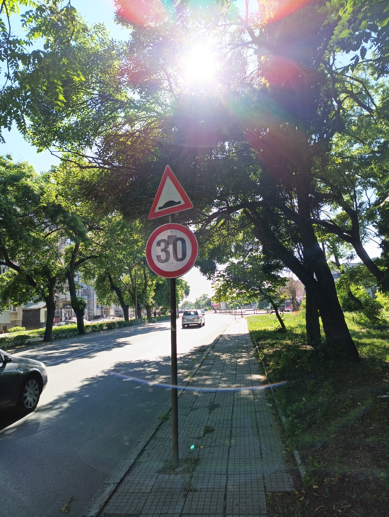 In the city of Karnobat, Bulgaria, on a bright, sunny day, a street scene is captured with a focus on traffic signs and the surrounding environment. The image shows a road with a sidewalk to the right. A traffic pole stands on the sidewalk, displaying two signs. The upper sign is a red-bordered triangle with a black depiction of a speed bump. Below it, a circular sign with a red border contains the number "30" in black, indicating a speed limit of 30 kilometers per hour.
The road is asphalted, and a portion of a dark-colored car is visible on the left side of the frame, driving on the road. Further down the road, a white car is seen moving away from the viewer. The sidewalk is paved with square tiles. Lush green trees with dense foliage line the street on both sides, casting shadows on the road and sidewalk, creating a dappled light effect. In the background, buildings with windows and balconies are visible behind the trees, suggesting a residential or commercial area. The sky is clear and blue, with bright sunlight causing lens flare and sunburst effects in the upper portion of the image. The overall atmosphere is that of a typical day in a moderately populated urban setting.