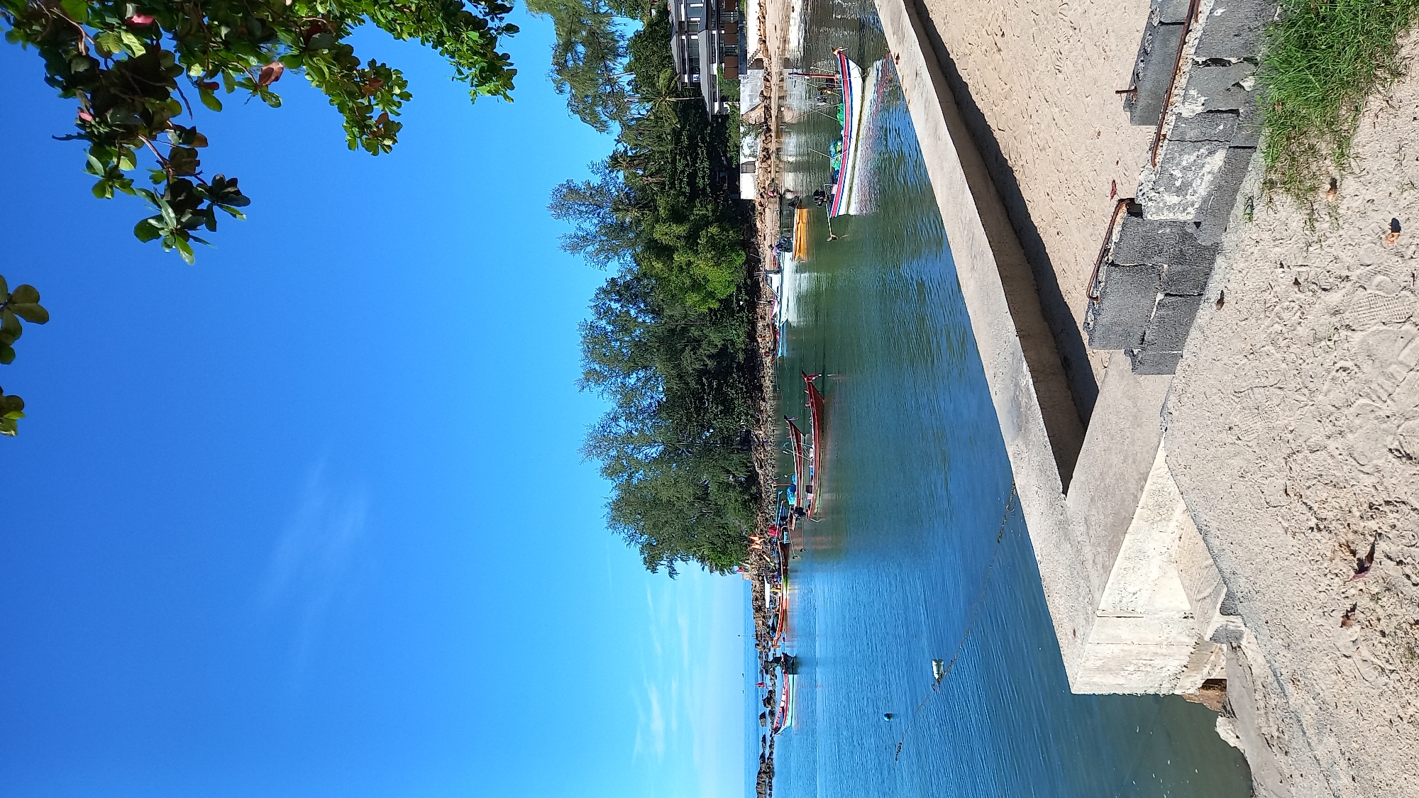 The image depicts a serene coastal scene in Ko Pha Ngan, Thailand, under a clear blue sky. The foreground features a sandy beach with a concrete structure, possibly a breakwater or a partially constructed seawall, with some loose concrete blocks visible. Beyond the sand, a calm body of turquoise water stretches out, dotted with numerous small, colorful fishing boats. These boats are moored and appear to be at rest. In the middle ground, a rocky coastline is visible, densely covered with lush green trees and vegetation. Further back, a modern building with a dark roof and white walls is nestled amongst the trees, suggesting a resort or residential area. The overall impression is one of peaceful tranquility and a typical coastal environment in a tropical location.